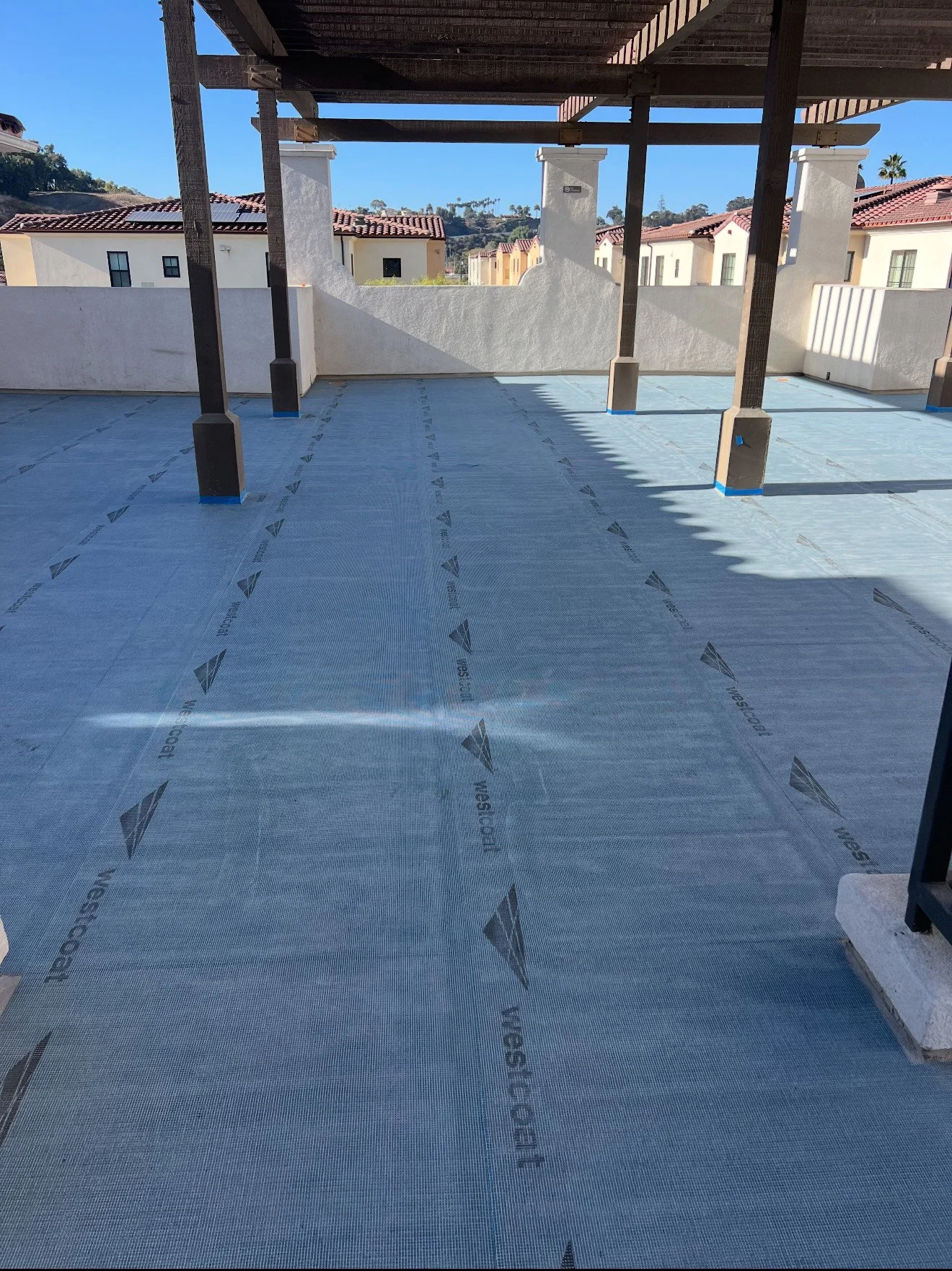 An outdoor balcony or terrace under construction with a blue weatherproof membrane on the flooring, supported by wooden beams and surrounded by a white wall, in a residential area with houses and hills in the background.