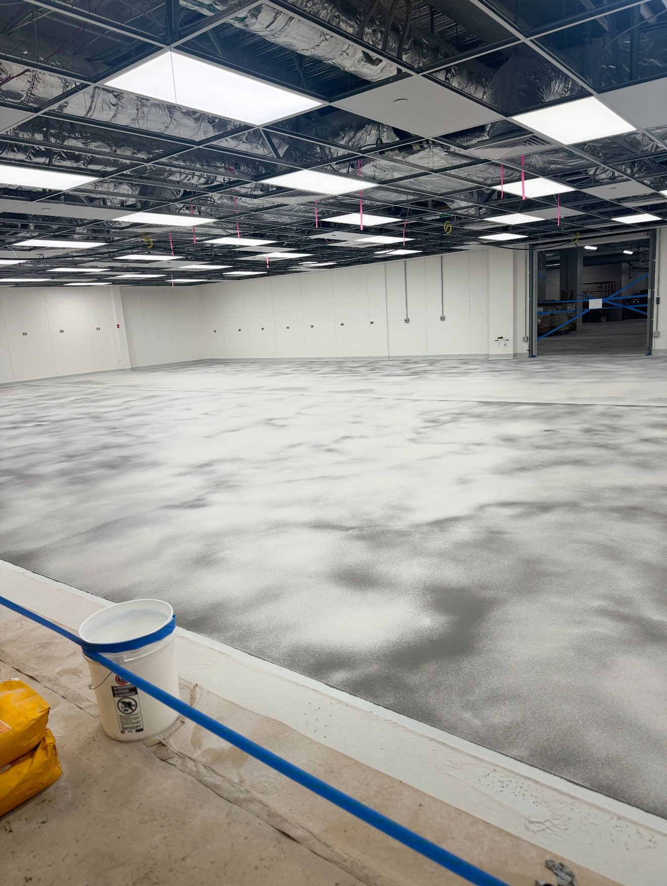 An unfinished indoor space under construction with exposed ceiling framework, drywall walls, and a concrete floor with a textured finish. There are construction materials and a plastic bucket in the foreground.
