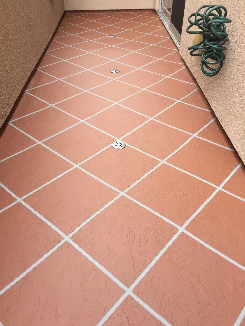 Terracotta-colored tiled outdoor patio with white grout lines, drain covers, a coiled green garden hose on the wall, and a sliding glass door.