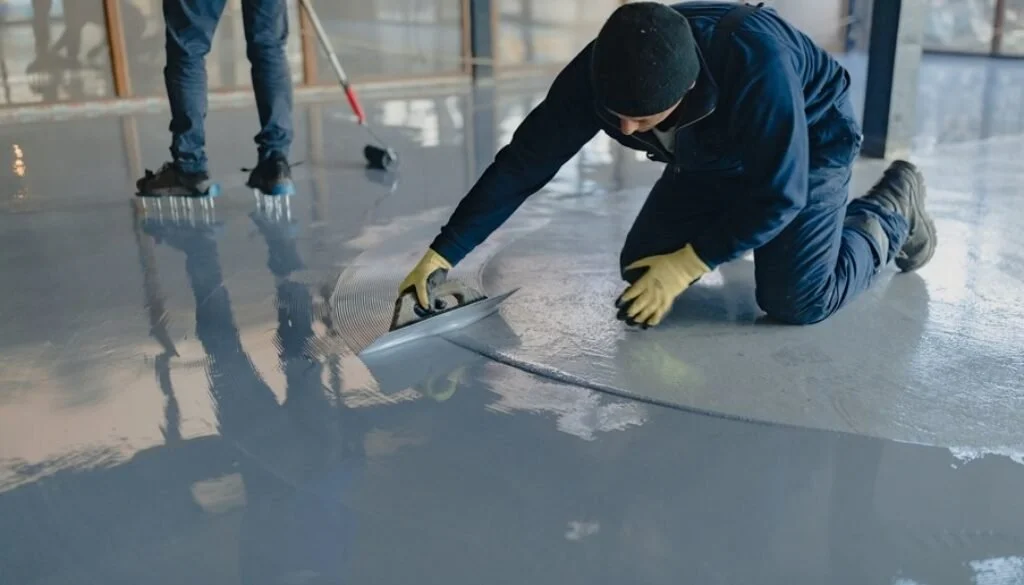 A worker in a black hat and jacket is kneeling and smoothing a freshly poured concrete floor using a trowel, with another worker in the background also working on the floor.