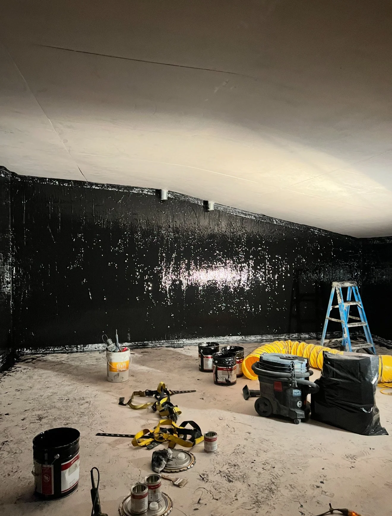 A room under renovation with black waterproofing paint on the walls, construction tools, cans of paint, a ladder, a vacuum, and ventilation ducting.