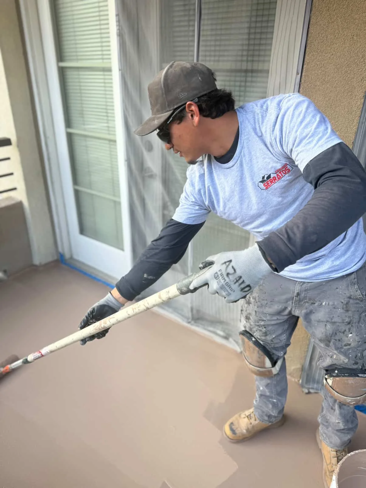 A worker in a gray shirt, dust-covered pants, and yellow shoes is applying a fresh coat of tan-colored liquid to a concrete porch using a long-handled roller.