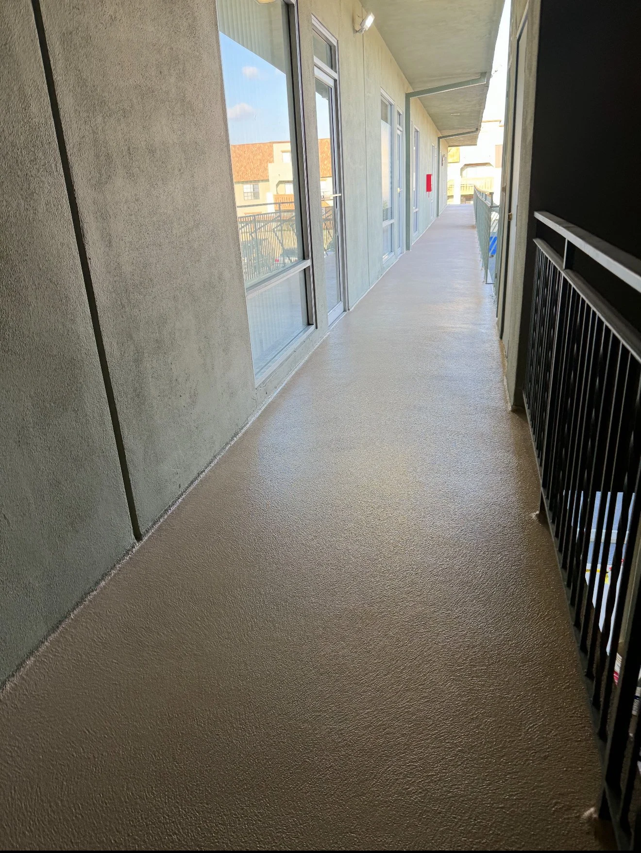 An outdoor corridor of an apartment building with beige textured flooring, green walls, glass doors, and black metal railings on the right.