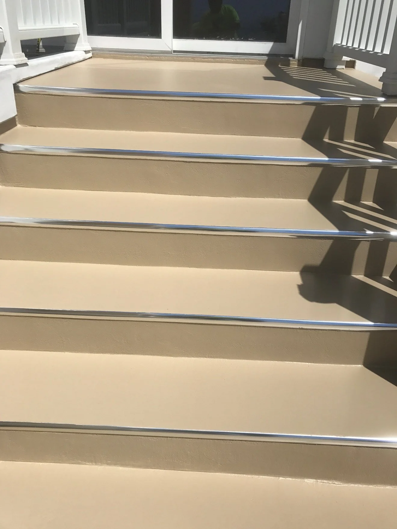 Exterior beige steps with silver handrails leading up to a sliding glass door, shedding shadows from a nearby railing.