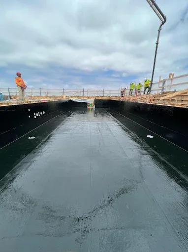 Construction workers on a rooftop under a cloudy sky, pouring black waterproof coating into a flat rooftop area.
