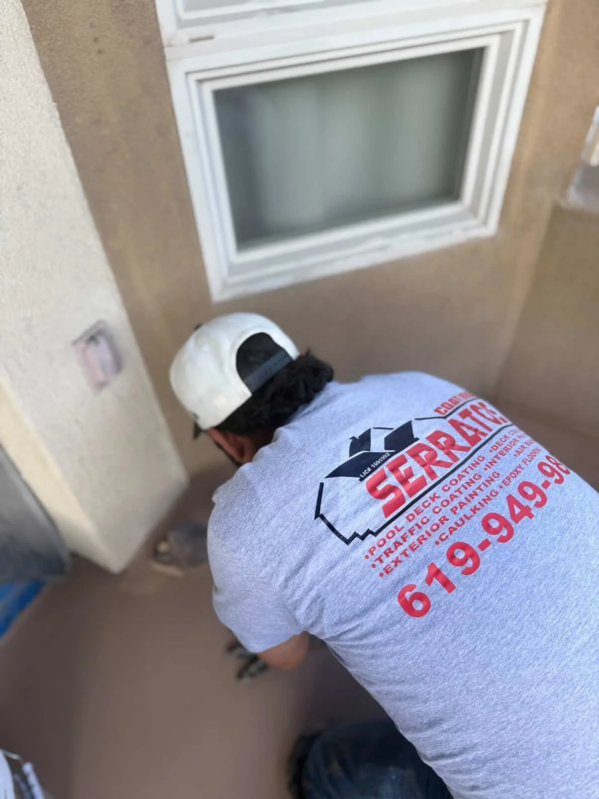 A worker wearing a beige cap and a gray T-shirt with red and black text is crouched down, working on a small outdoor area near a window on a beige wall.