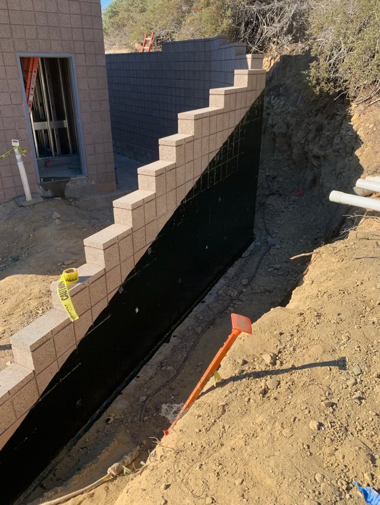 Construction site with a partially built block wall, a doorway opening, and a trench with a black waterproofing barrier, construction tools, and caution tape.