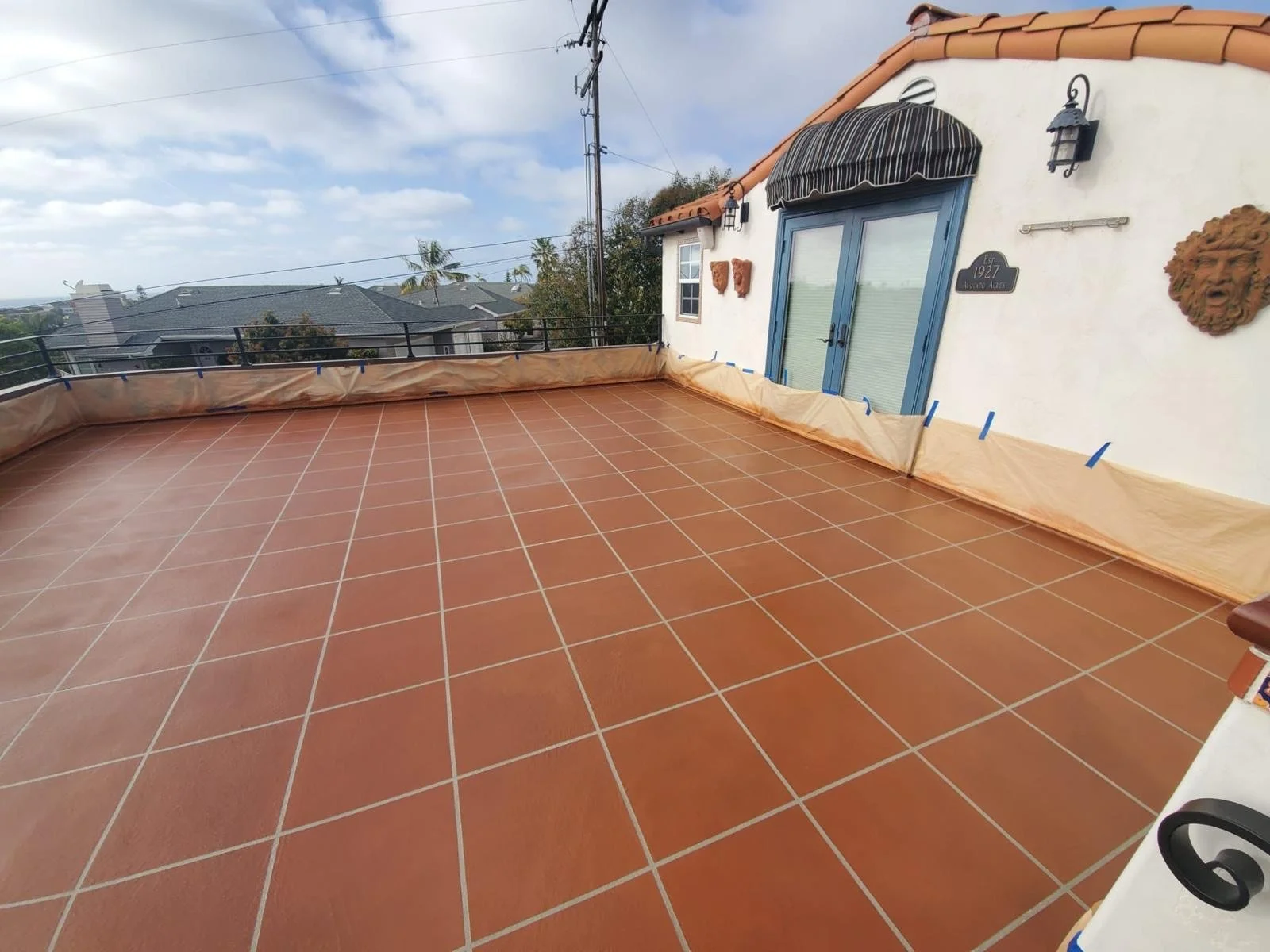 A rooftop terrace with terracotta tile flooring, a blue door with a striped awning, decorative wall sculptures, and a cloudy sky in the background.