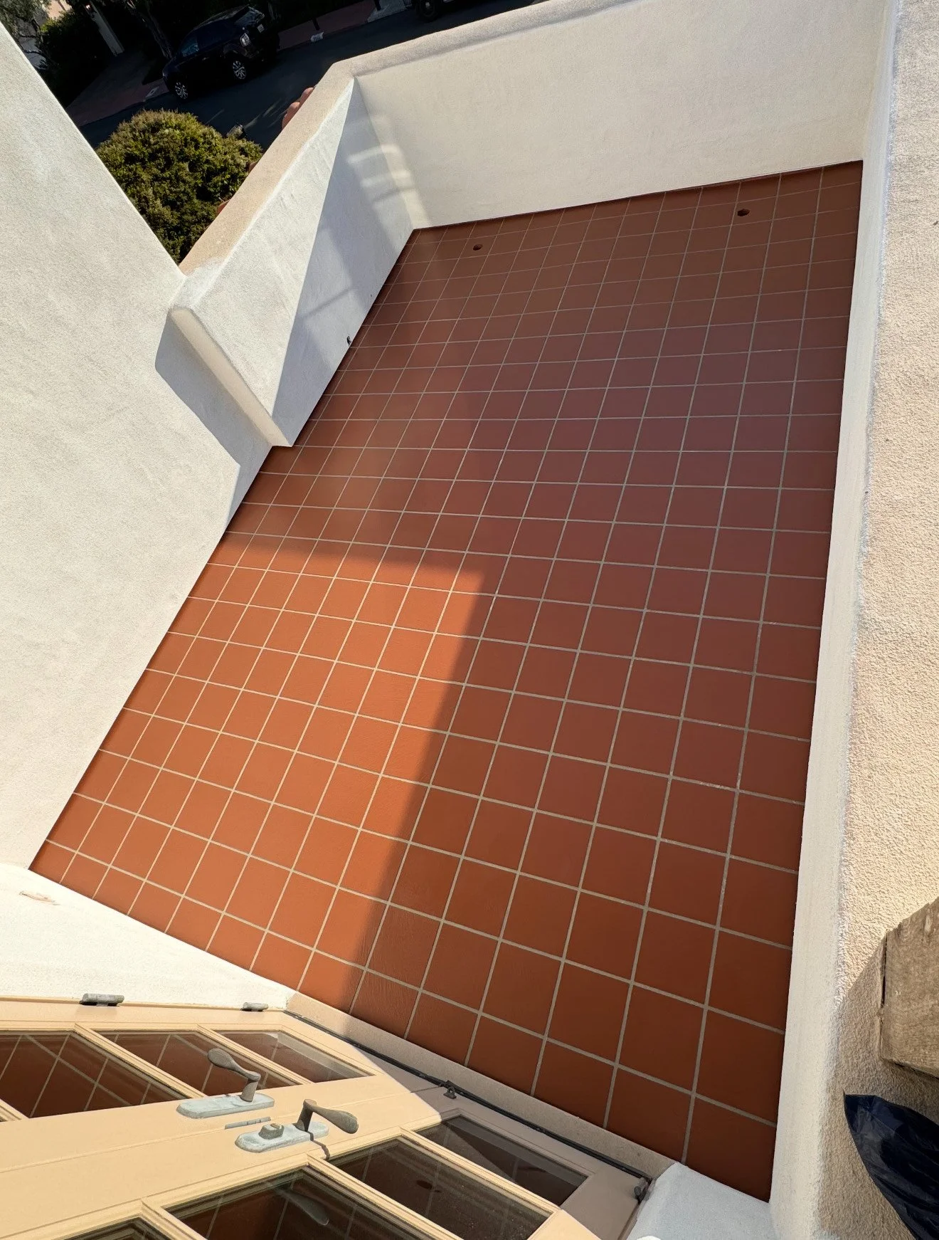 A small balcony with terracotta-colored tiled floor, white surrounding walls, and a beige door with glass panels at the bottom of the image. Outside is a parking lot with a few cars and a bush.