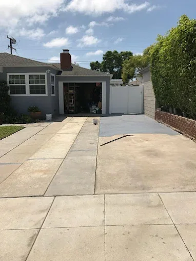 Residential driveway leading to a garage with an open door and a partly paved section, surrounded by a house with a chimney and a green hedge on the right.