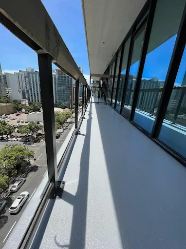 View of a balcony with glass railings overlooking a cityscape with tall buildings, parked cars, and trees, under a clear blue sky.