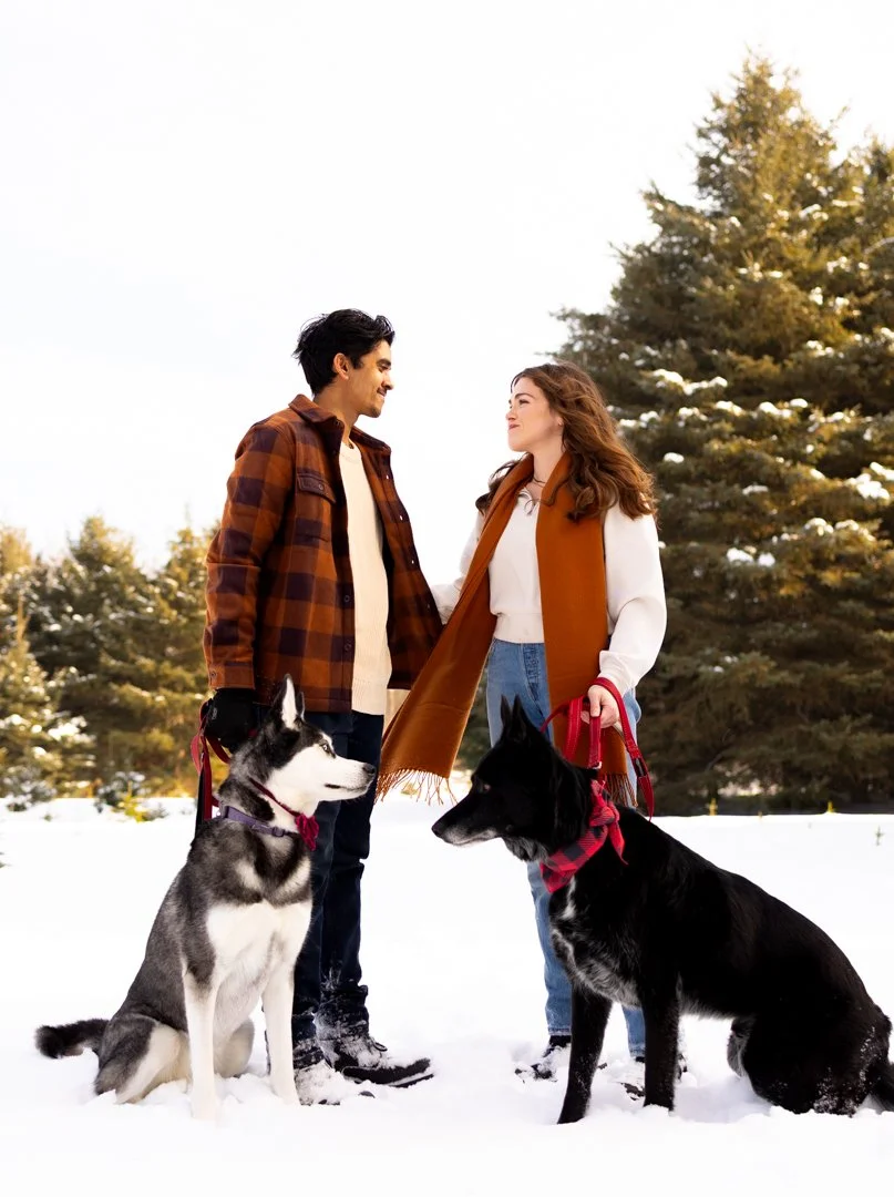 A young couple standing in the snow with two dogs, looking at each other and smiling, in a winter landscape with snow-covered trees.