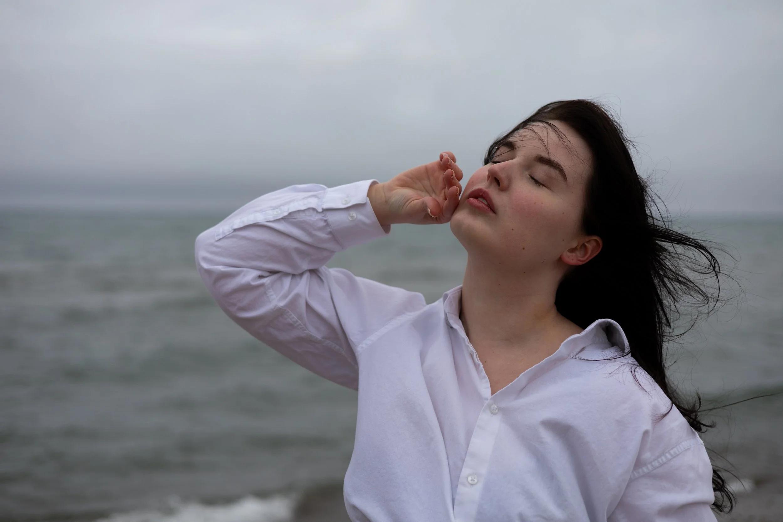 A woman in a white shirt standing by the ocean on a cloudy day, with her eyes closed and her hand touching her face, wind blowing her hair.