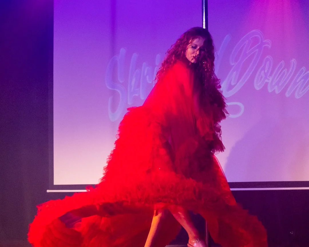 A woman with curly hair wearing a long, flowing red dress performing on stage, with purple lighting and a backdrop with faint writing.