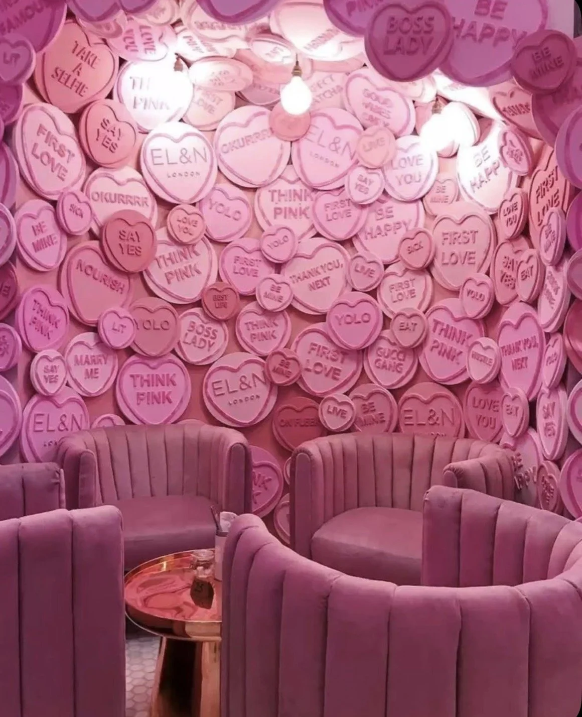 Pink themed room decorated with numerous pink heart-shaped pillows with various love and romantic messages, surrounded by pink velvet armchairs and a small round table.