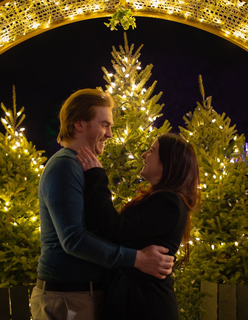A couple smiling and embracing near decorated Christmas trees with lights, under an illuminated arch at night.