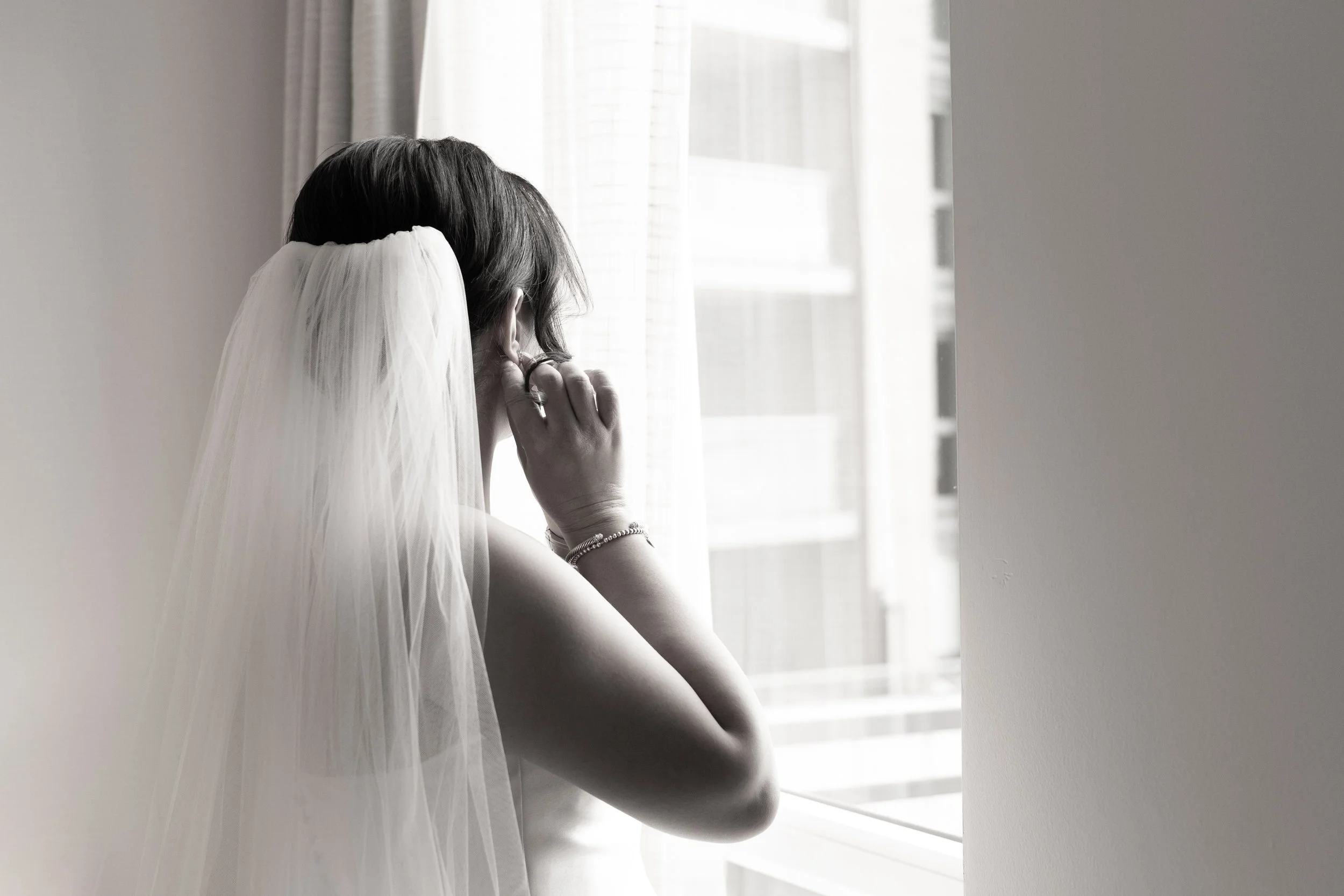 A bride wearing a veil and earrings gazes out of a window.