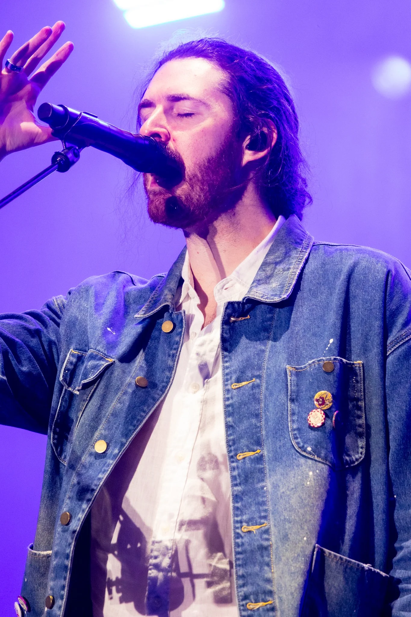 A man with long hair and a beard singing into a microphone, wearing a denim jacket and a white shirt, with purple lighting in the background.