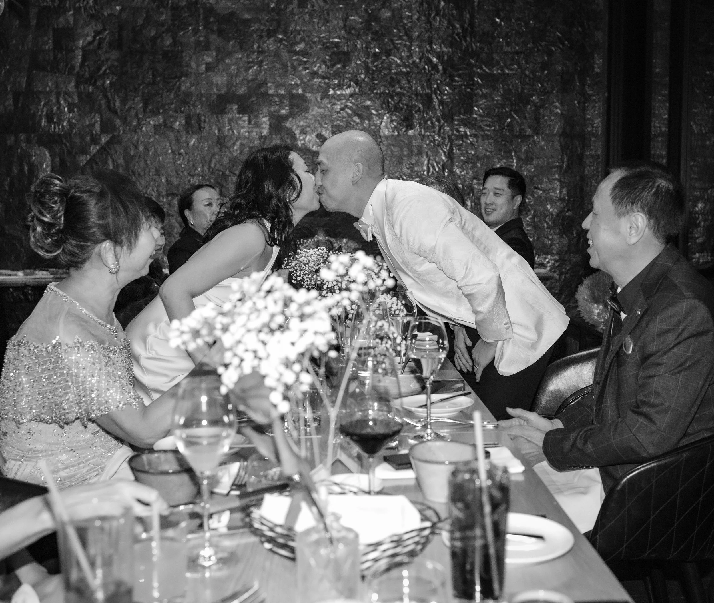 A black and white photo of a group of people at a formal dinner. Two people are leaning over the table and kissing. Others are seated and smiling, with glasses of wine and floral centerpieces on the table.