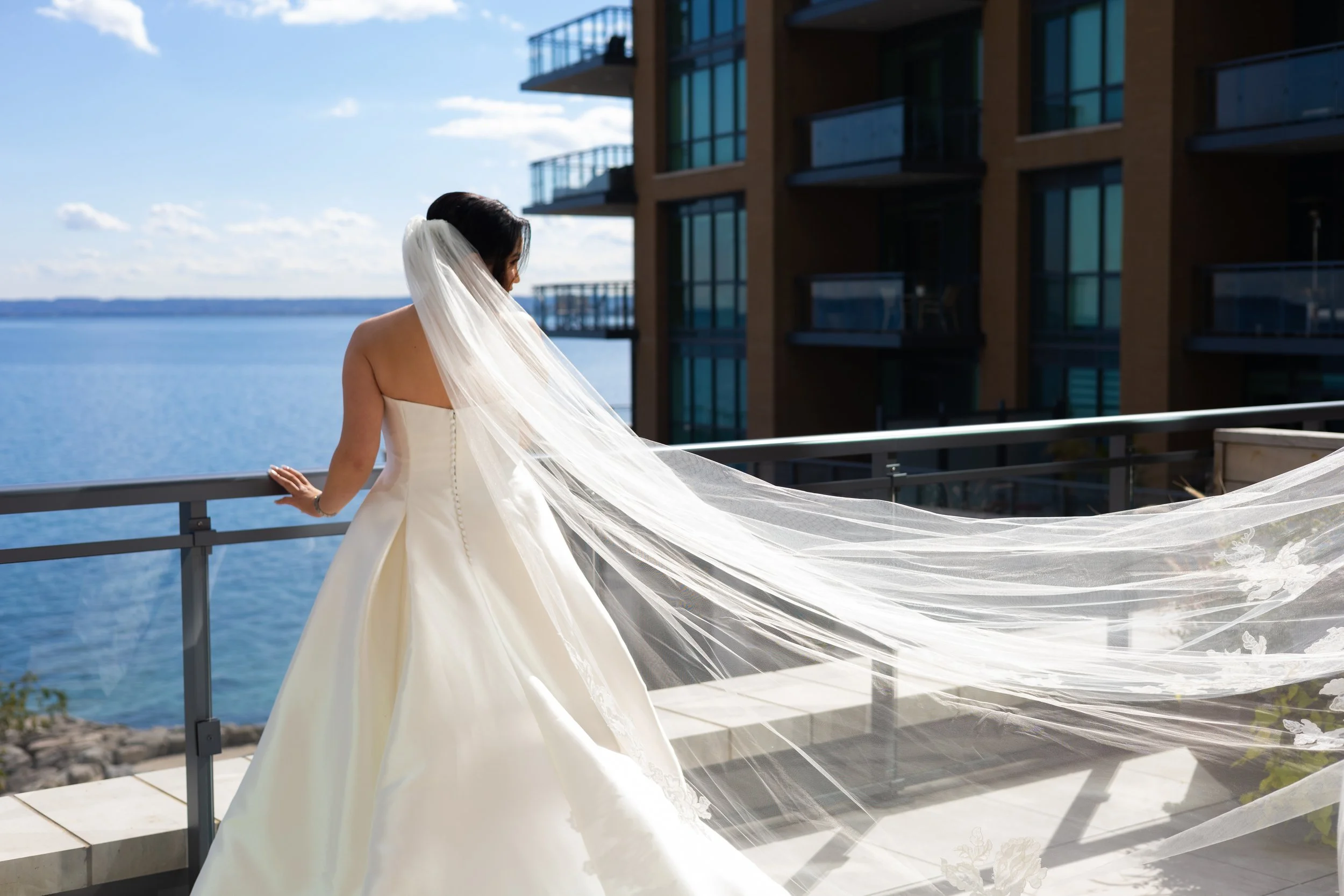 Bride in white wedding dress with long veil standing on balcony overlooking water and modern buildings.