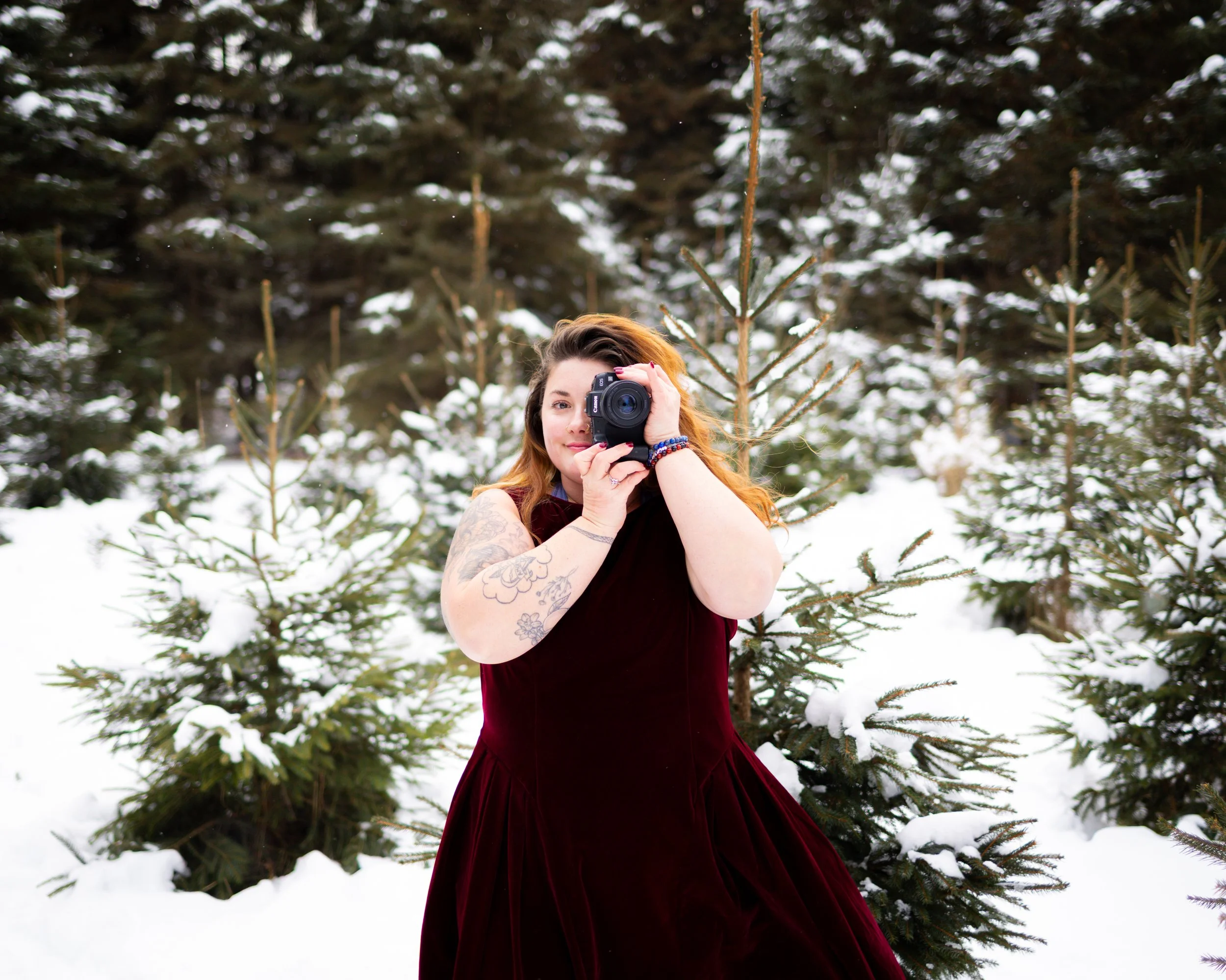 A woman in a red velvet dress holding a camera, standing in a snowy forest with evergreen trees and snow-covered ground.