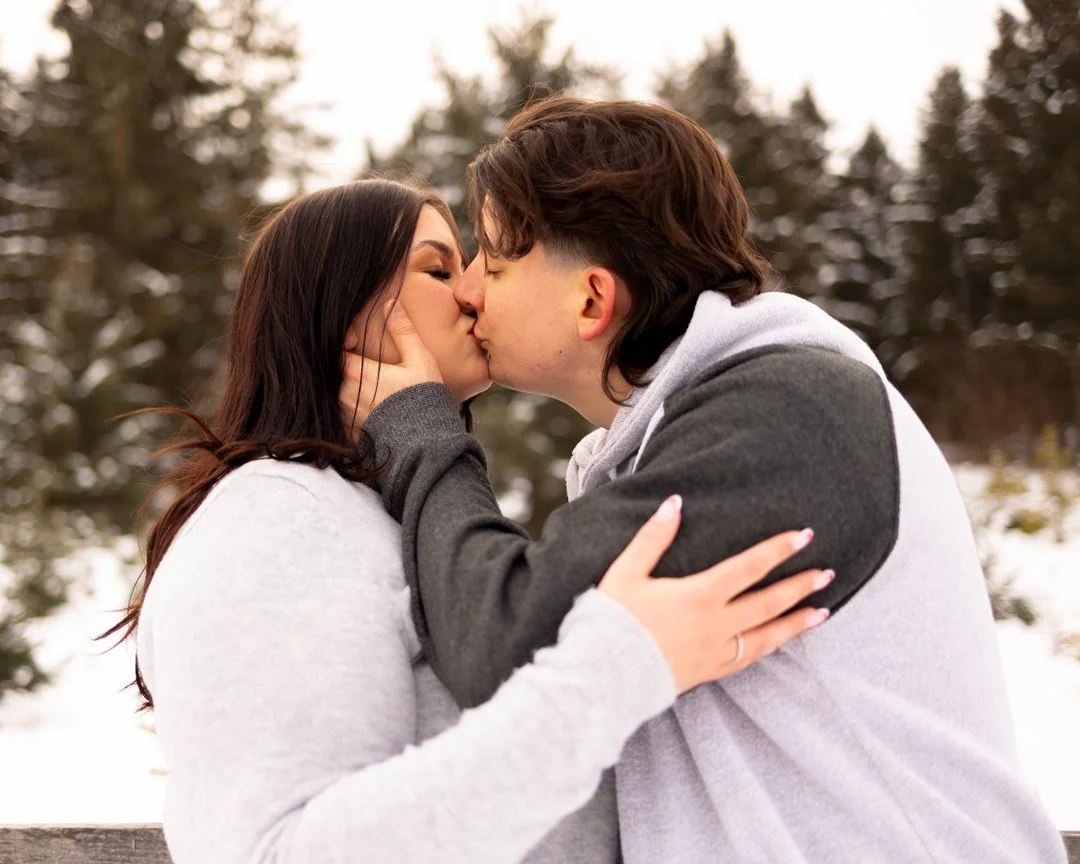 A couple kissing outdoors in winter, with snow and evergreen trees in the background.