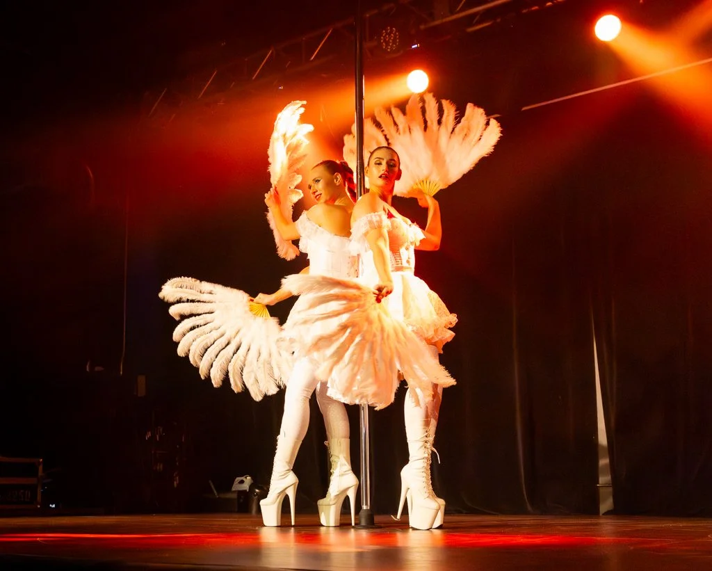 Two women perform a dance on stage with feather fans, wearing white costumes and high heels, under orange stage lighting.