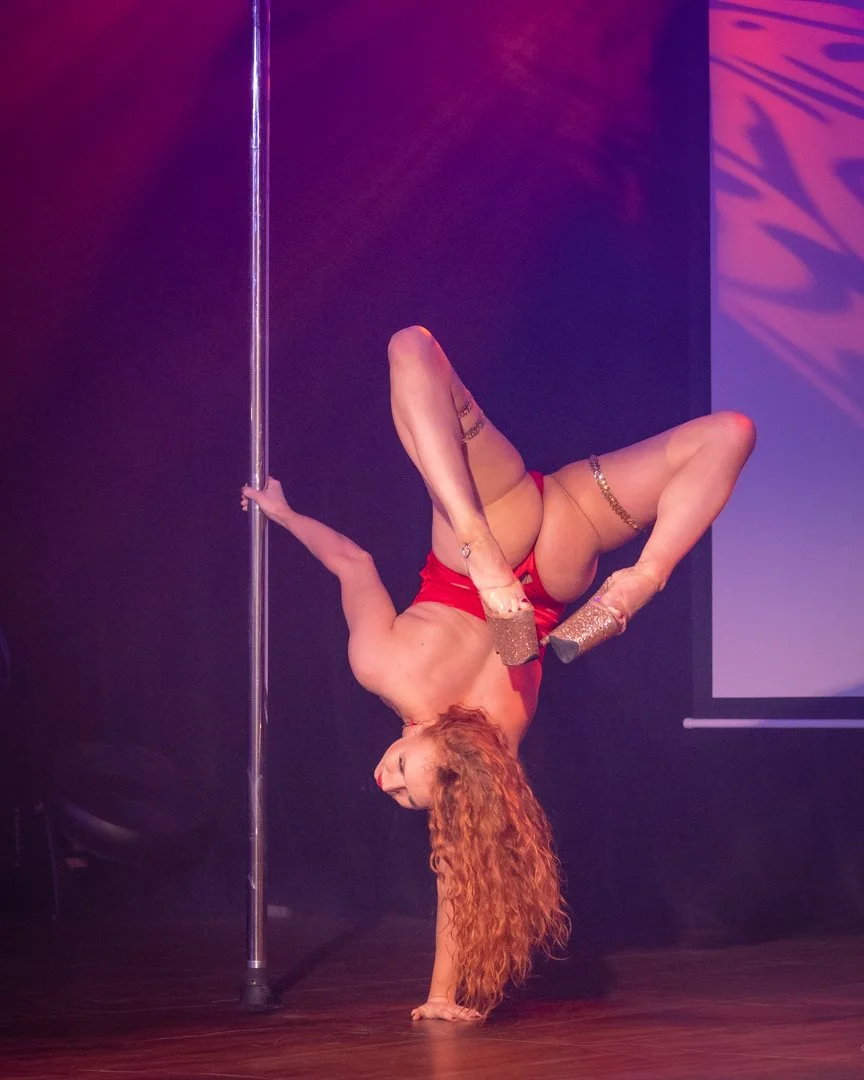 A woman with long, curly red hair performing a pole dance on a stage, wearing a red outfit and glittery shoes, illuminated by pink and purple lighting.