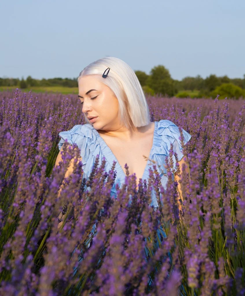 A woman with platinum blonde hair and a black hair clip standing in a field of purple lavender flowers, wearing a ruffled blue dress, with her eyes closed and facing slightly downward.