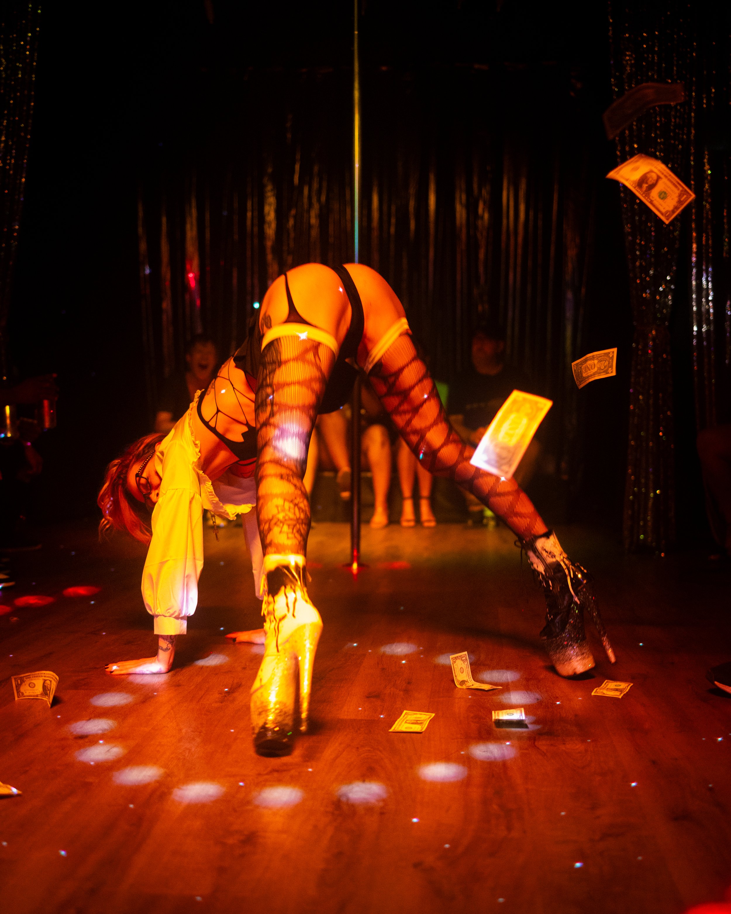 A woman in fishnet stockings, black lingerie, and gold high heels performing a pole dance on a wooden floor at a nightclub with dollar bills falling around her. She is in a backbend position with her hands and chest on the ground, and her hips raised