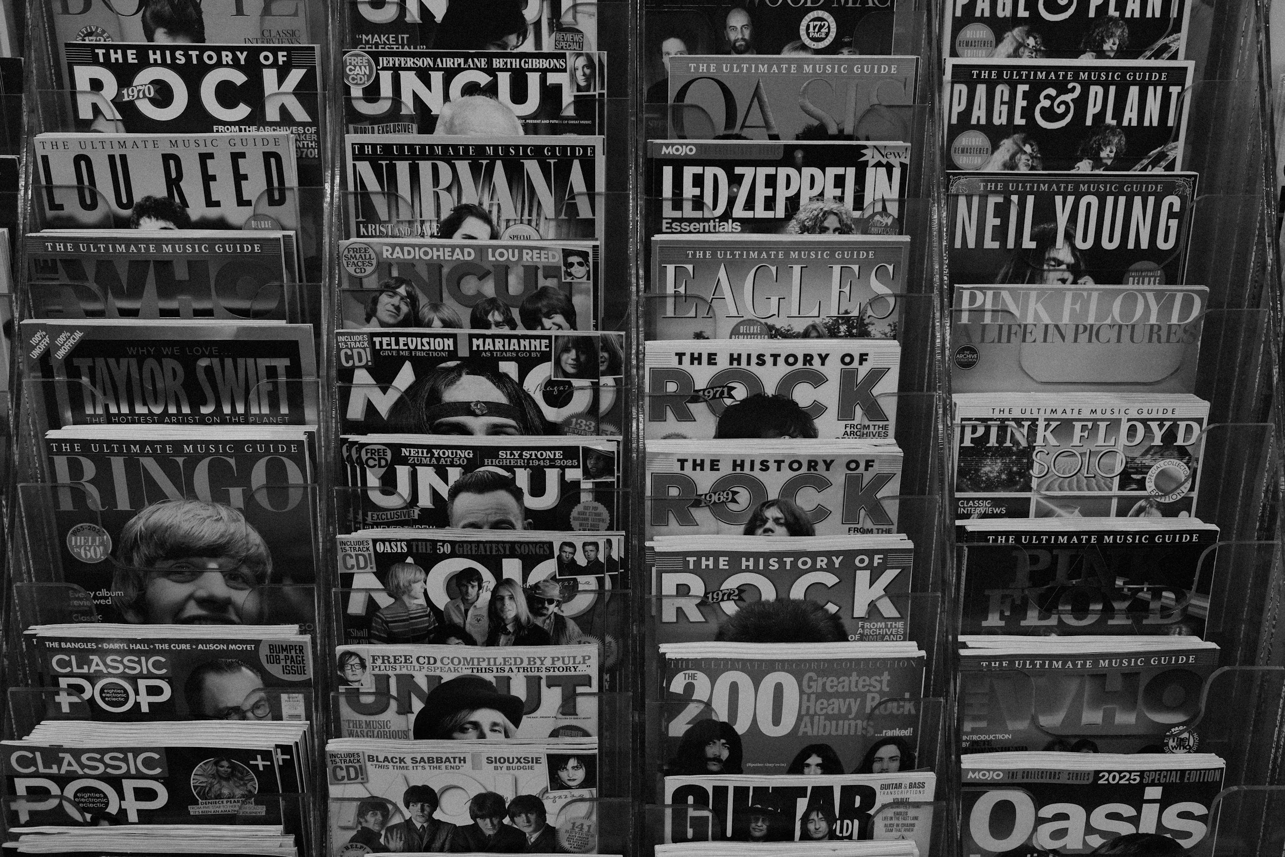 Black and white photo of magazine racks filled with music magazines and books on rock music, bands, and artists, with covers displaying images of musicians and bands such as Nirvana, Led Zeppelin, Pink Floyd, and Taylor Swift.