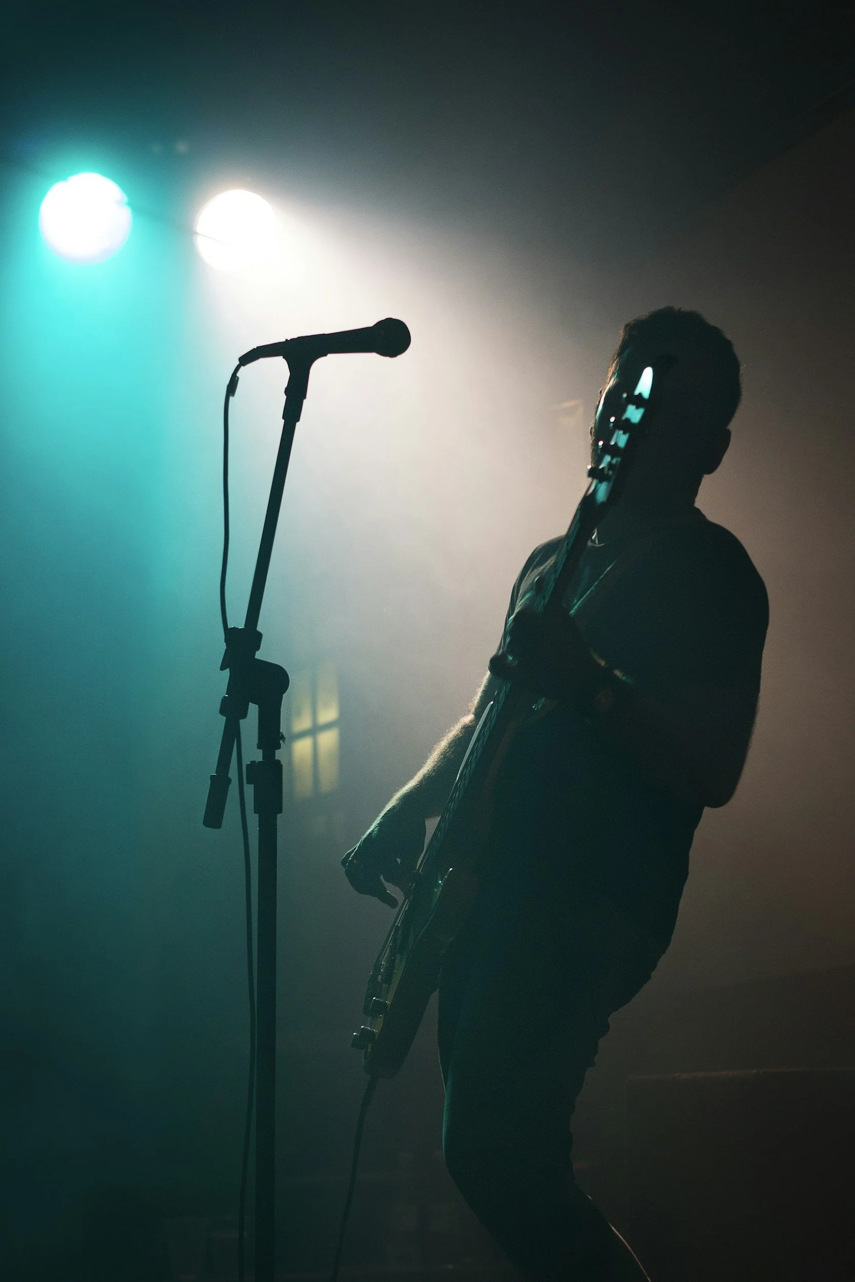 Silhouette of a musician holding a guitar next to a microphone on a dimly lit stage with blue and white lights.