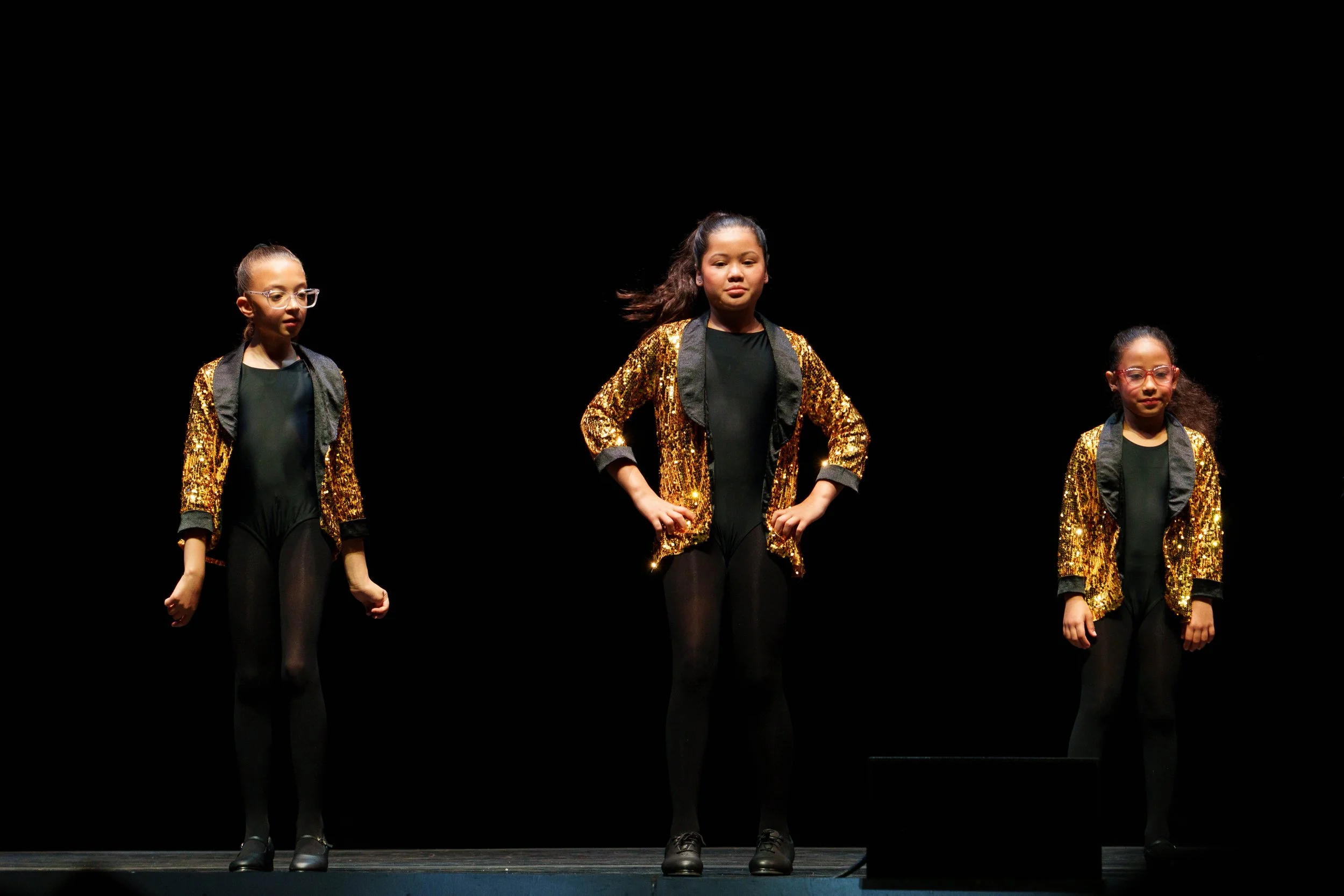 Three young girls performing on stage, wearing black leotards, black tights, and gold sequin jackets, with a dark background.
