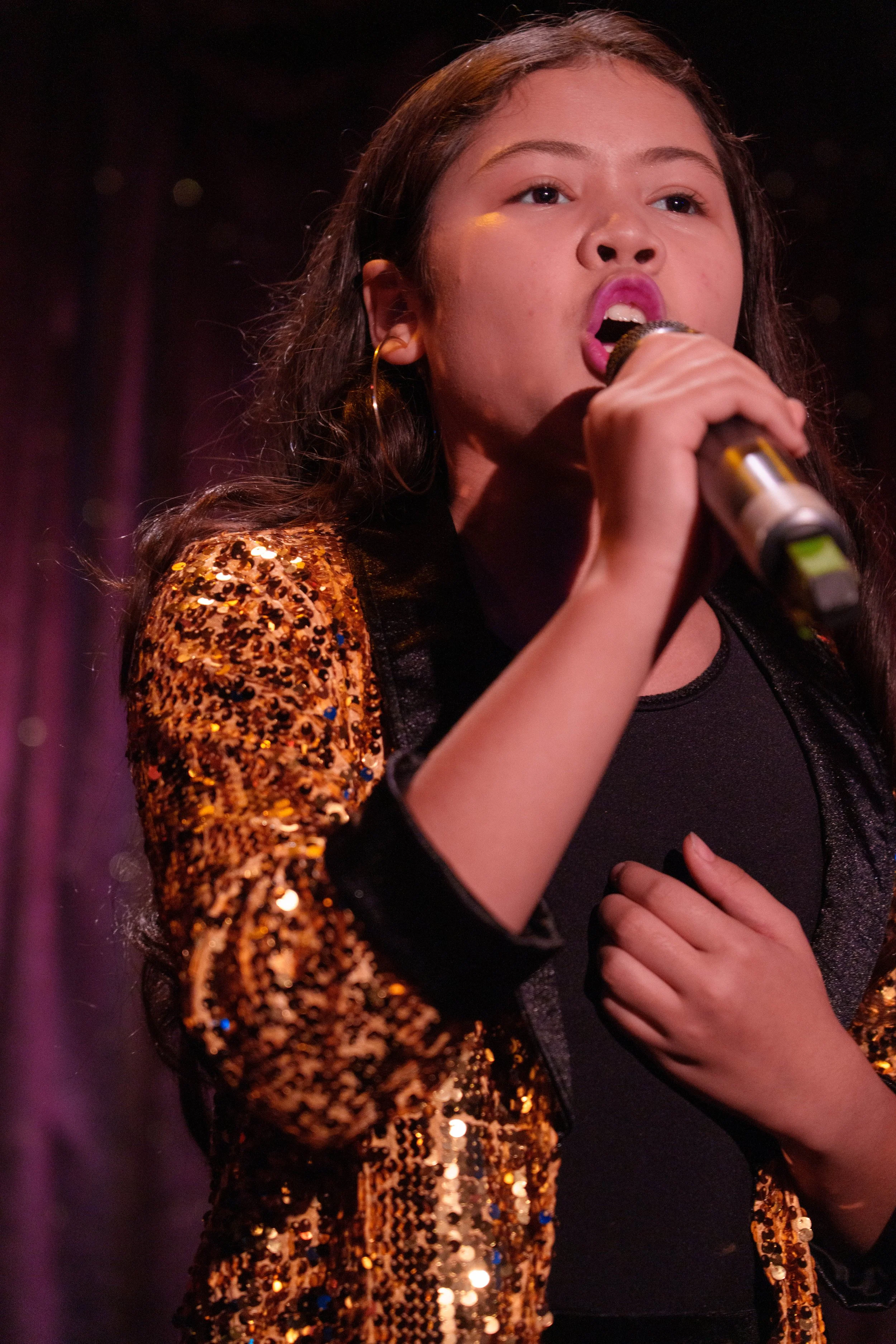A young woman singing into a microphone on stage, wearing a sequined jacket with black and orange colors.