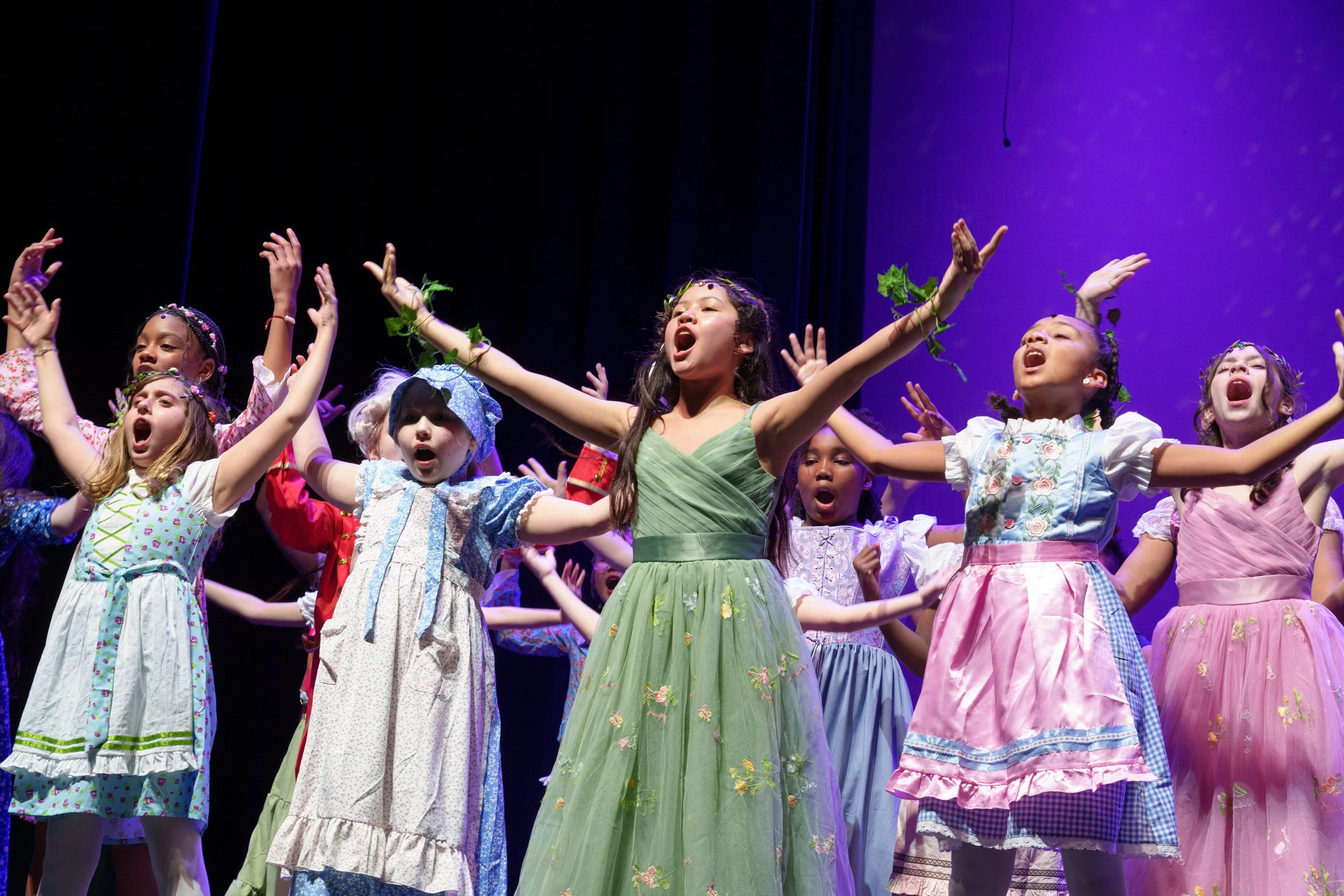 Children in colorful costumes perform on stage with arms raised and open mouths.
