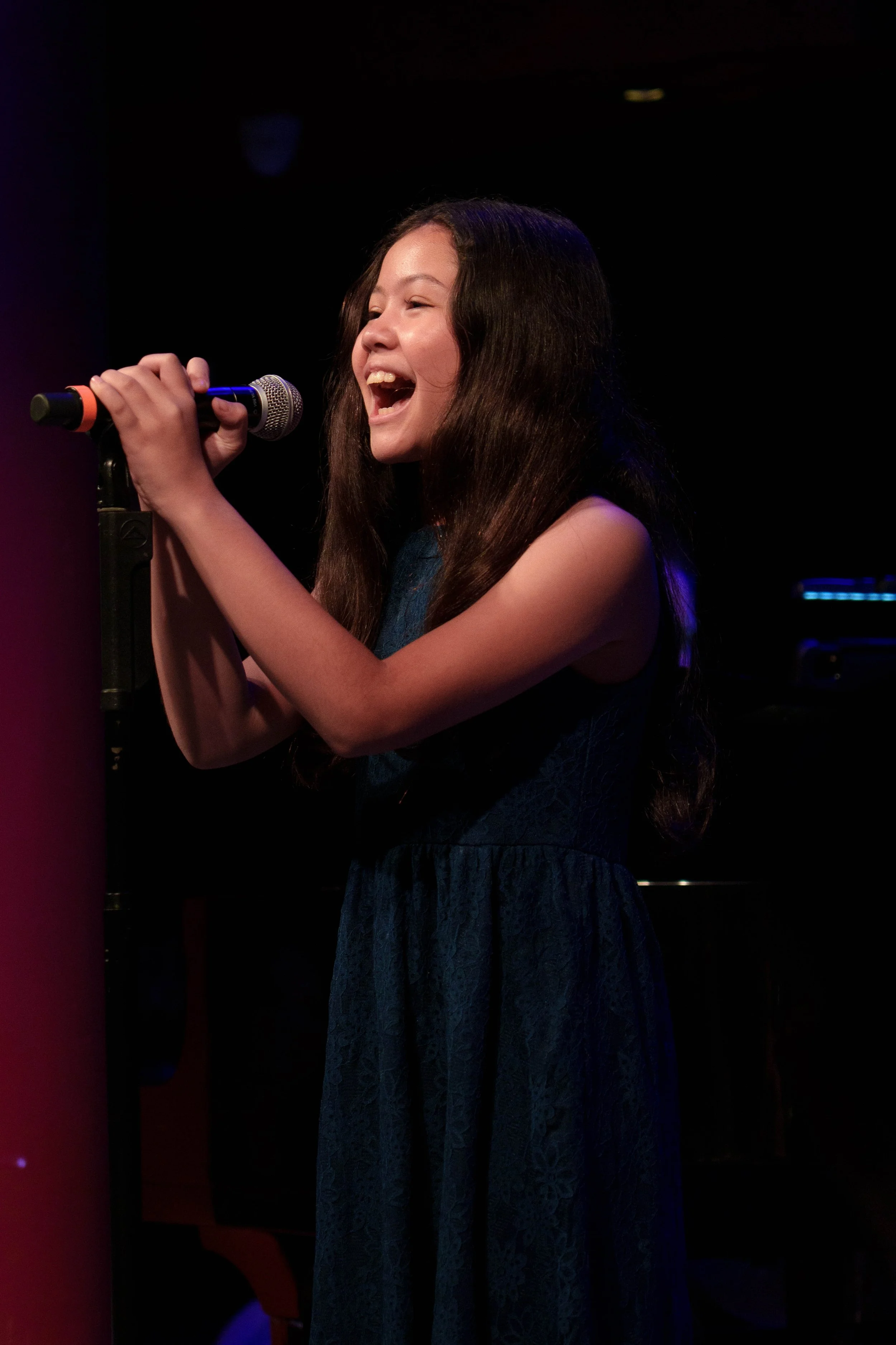 Young girl singing into a microphone on stage, wearing a dark blue dress, smiling and performing passionately.