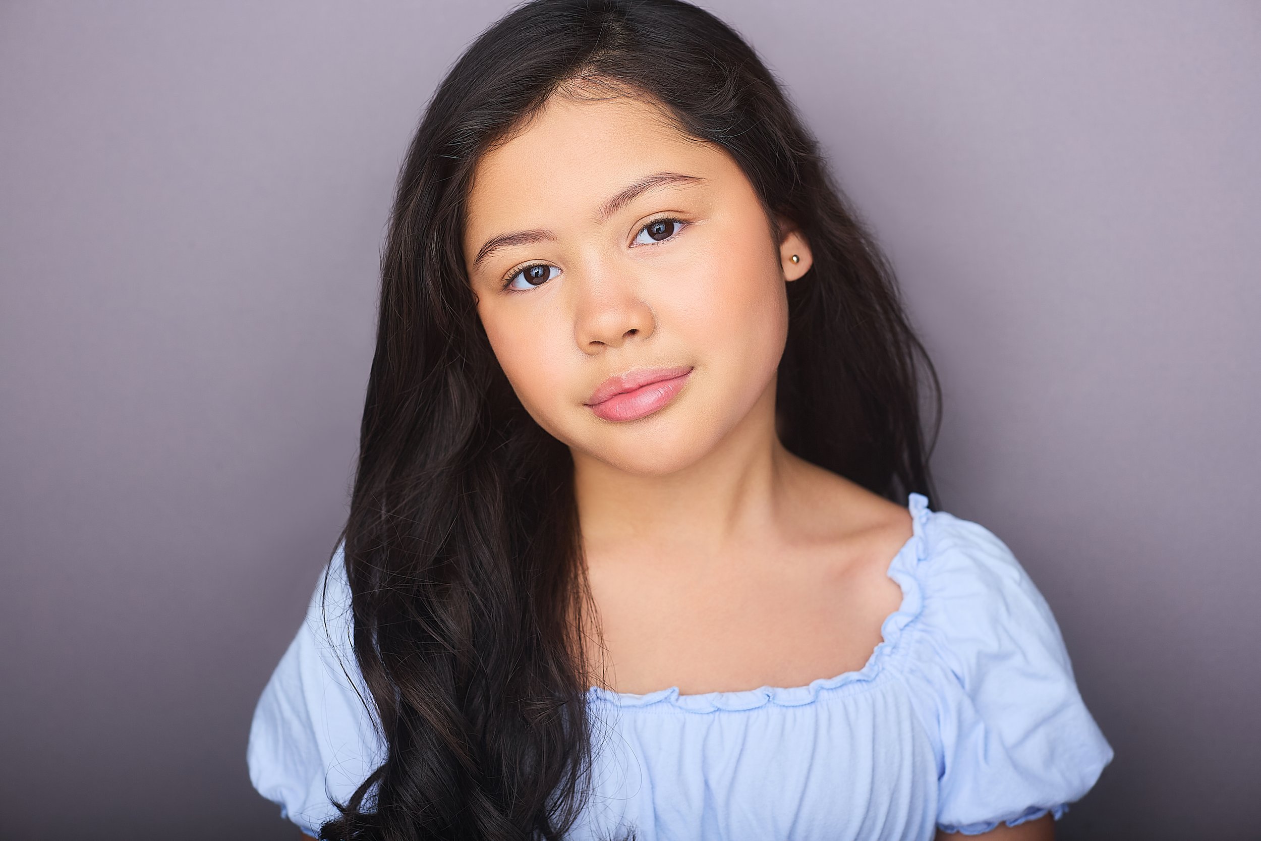 A young woman with long, dark wavy hair and light skin, wearing a light blue top with ruffled edges, standing against a plain gray background.