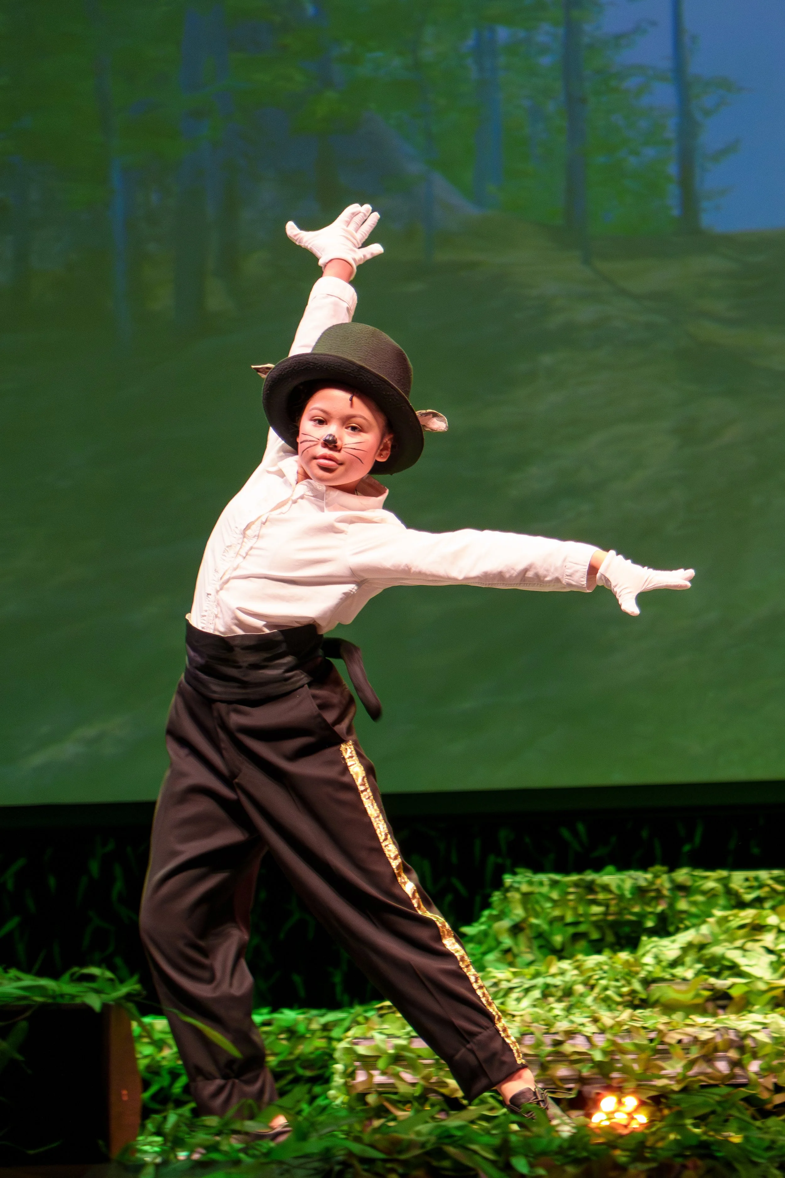 Child performing a jungle animal costume dance on stage, dressed as a mouse with a black hat, whiskers face paint, and light-colored gloves, against a backdrop of greenery.