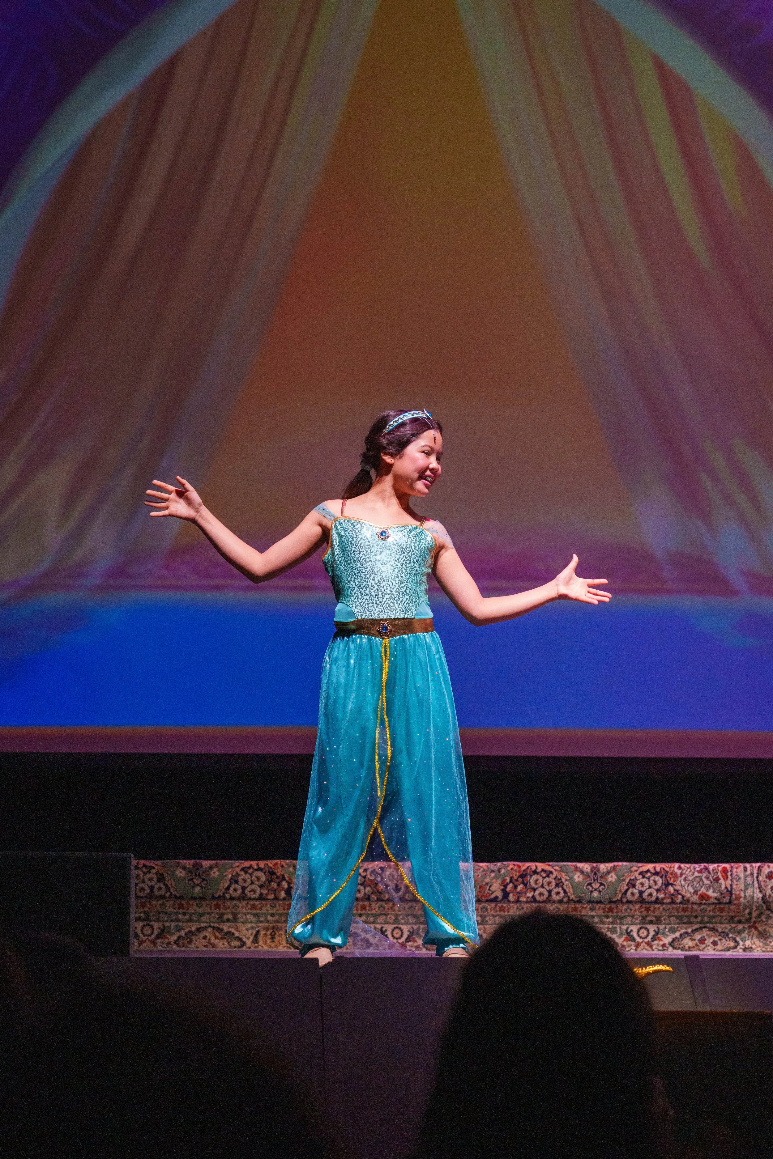 A young girl in a blue princess costume performing on stage with a colorful backdrop.