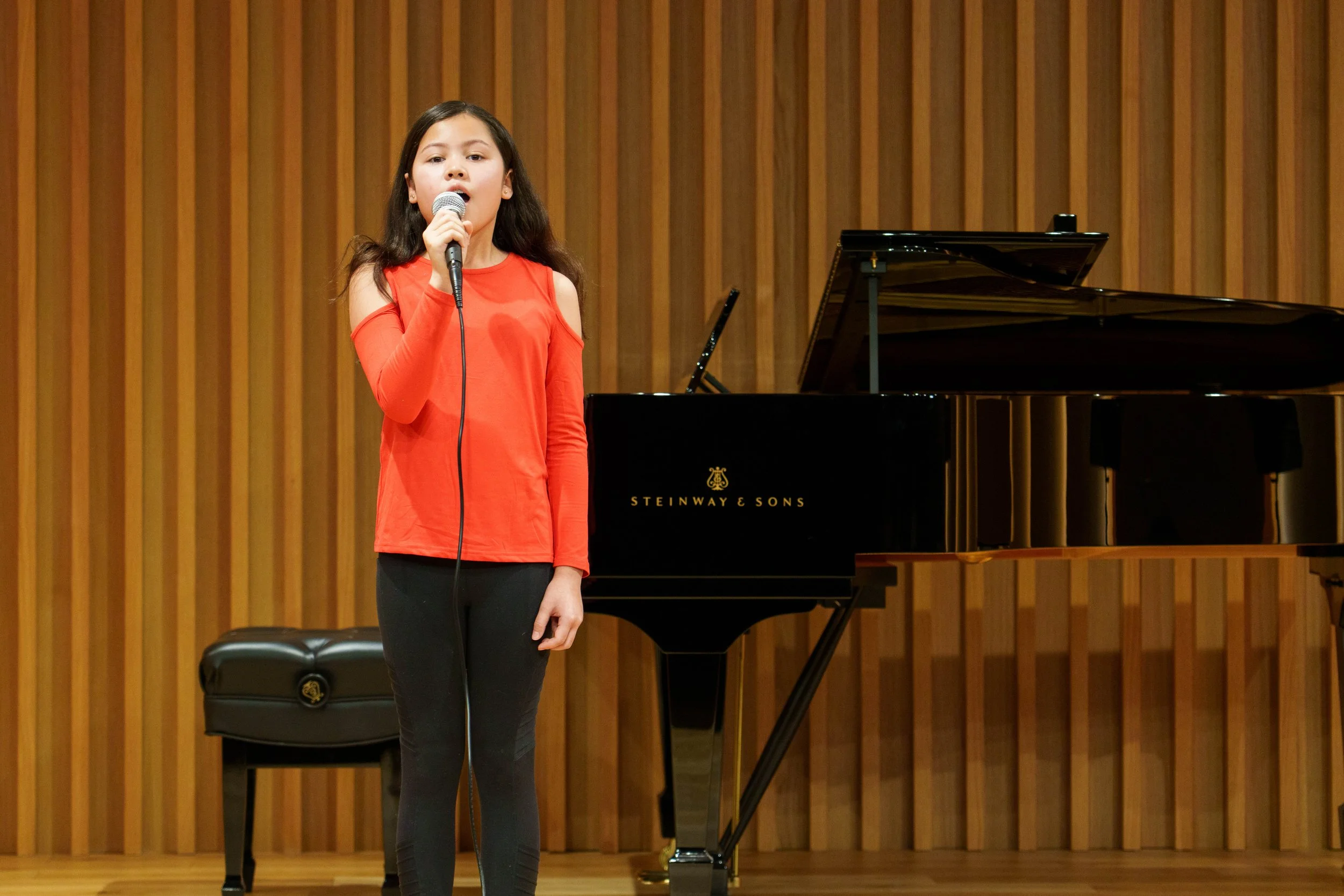 Young girl singing into a microphone on stage with a black grand piano behind her, wooden panel backdrop, wearing a red long-sleeve top and black pants.