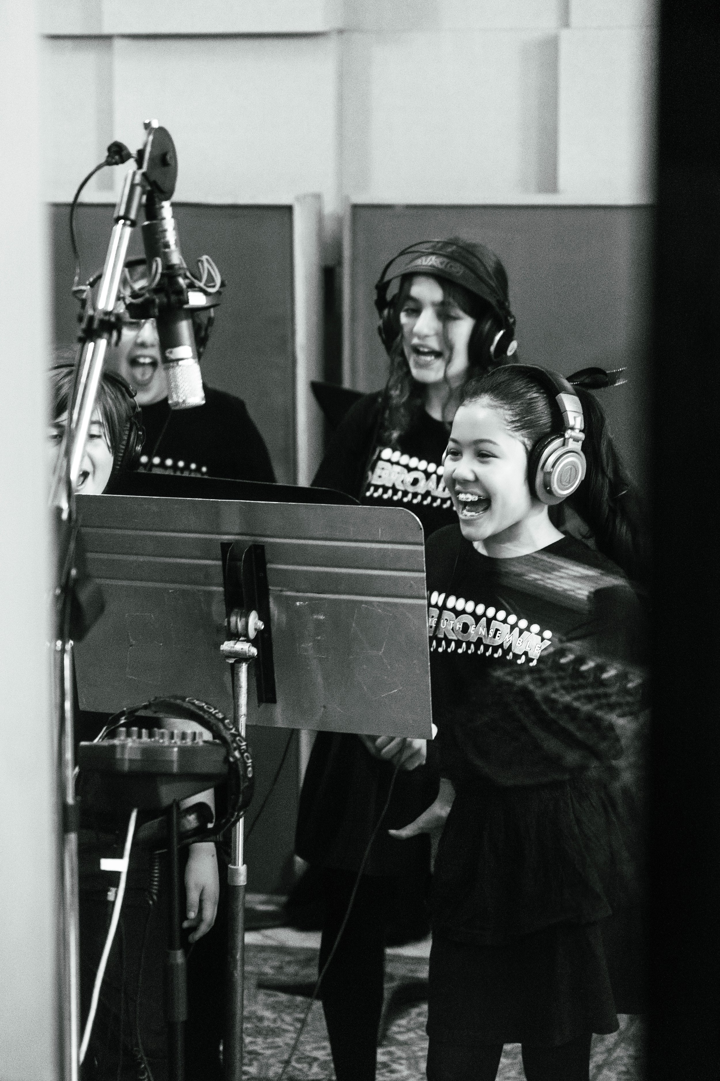 Group of children singing in a recording studio, wearing headphones and standing around a music stand.