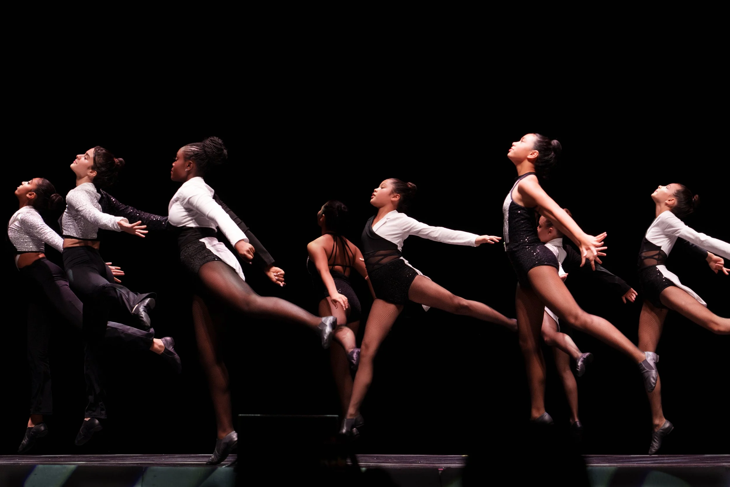 Group of dancers on stage performing a choreographed dance, dressed in coordinated black and white costumes, against a black background.