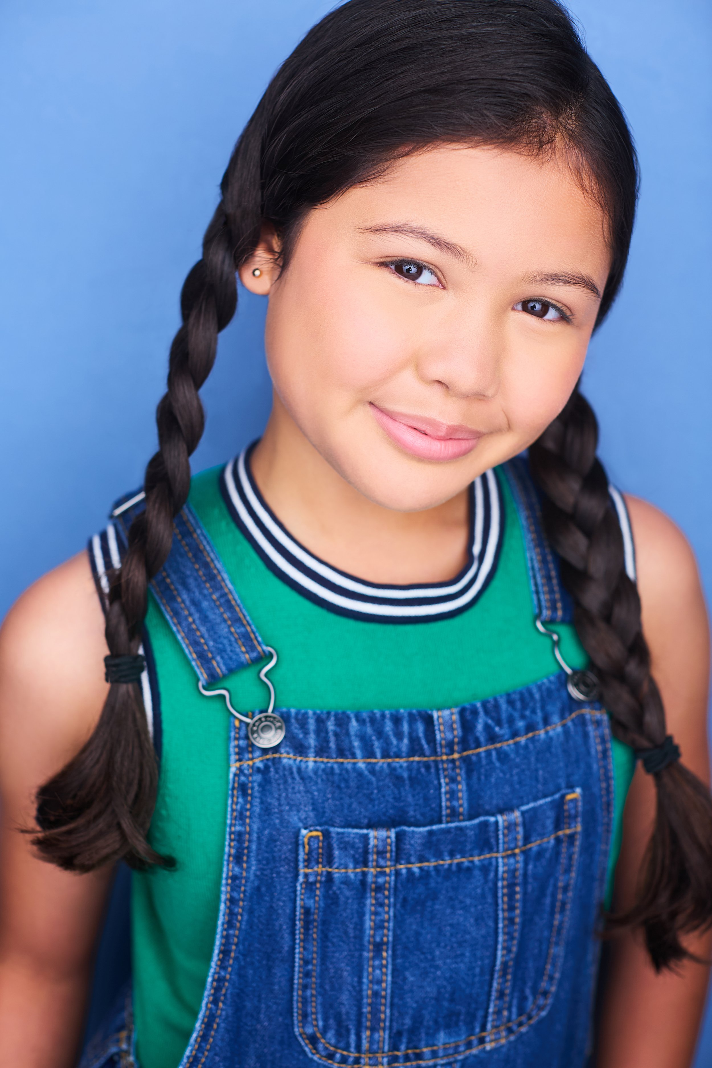 Smiling young girl with long braided hair wearing a green shirt and denim overalls against a blue background.