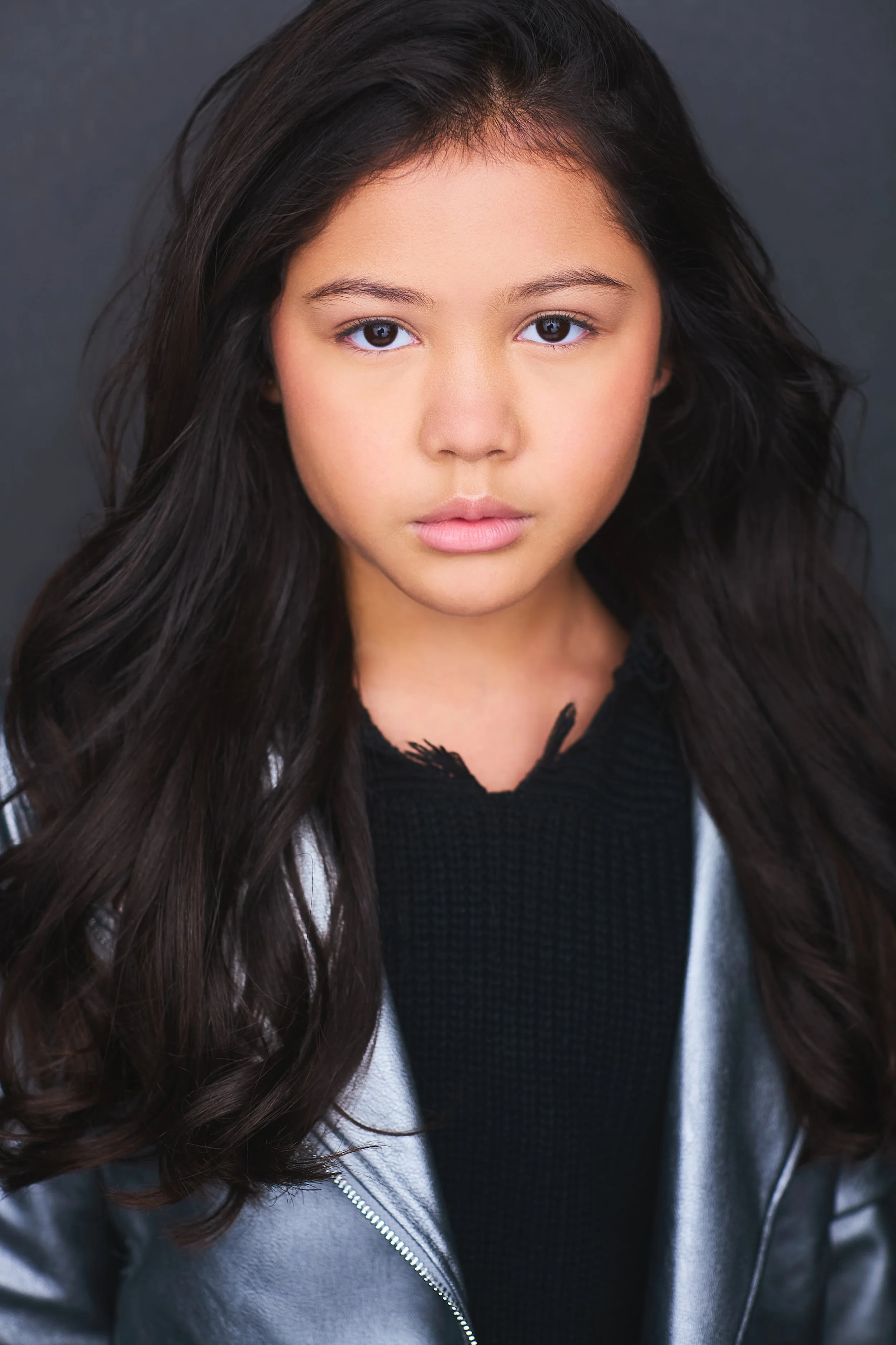 Close-up portrait of a young girl with long dark wavy hair, wearing a silver jacket over a black top, against a dark background.