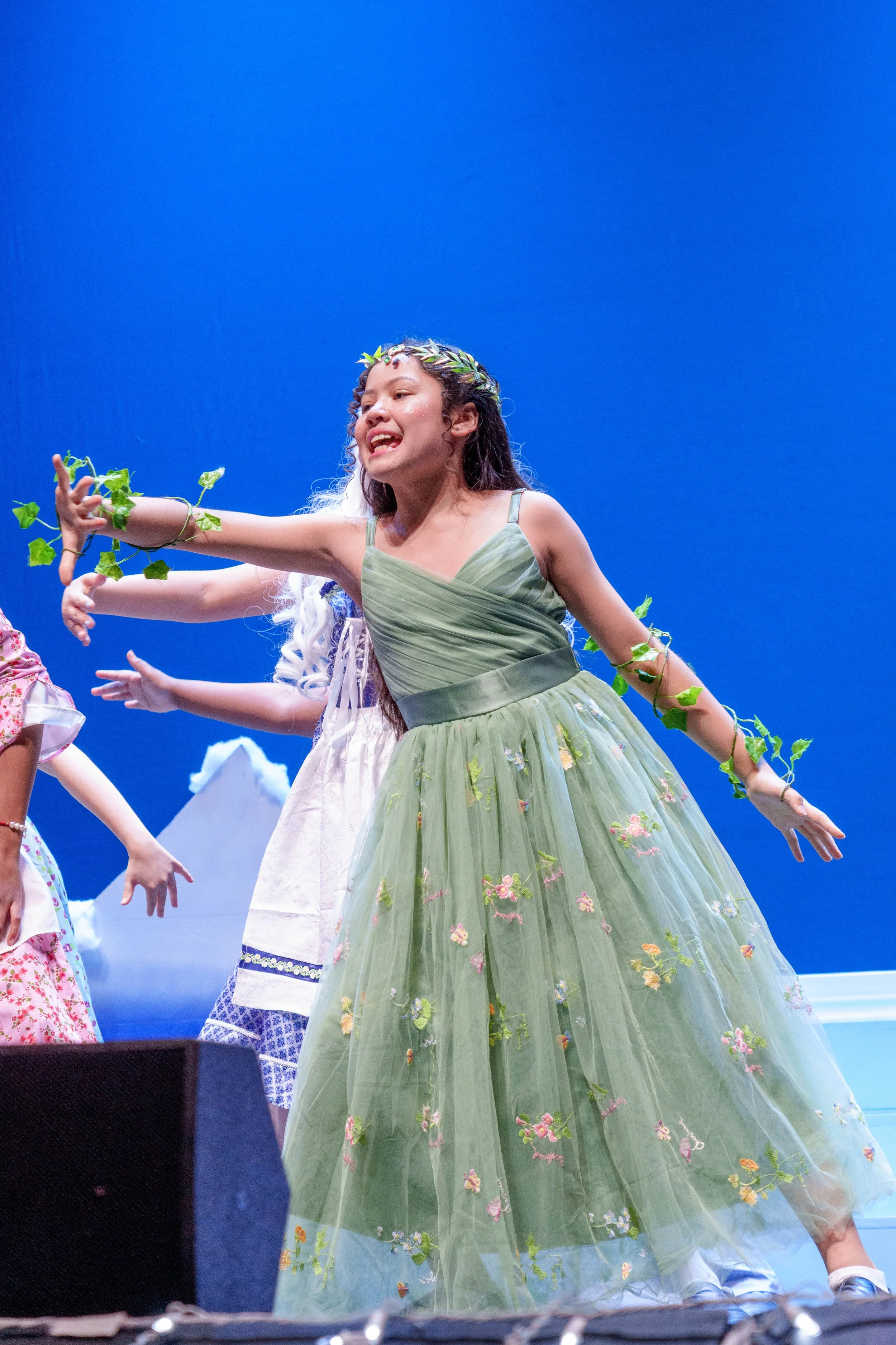Young girl dressed as a fairy or princess in a green floral dress, wearing a leafy crown and vine jewelry, performing on stage with a blue background.