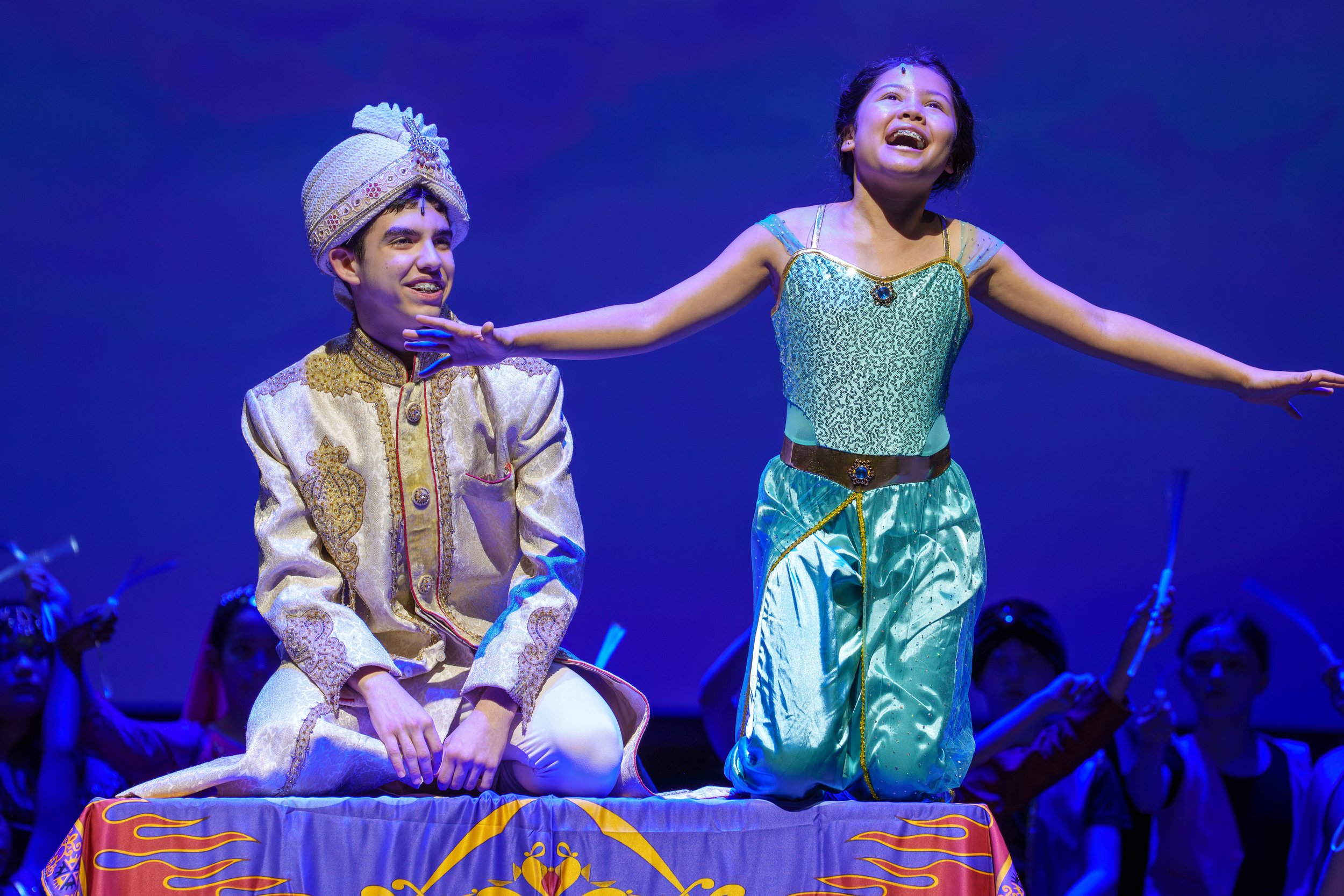 Young performers dressed as Aladdin and Jasmine from a stage play, with Jasmine extending her arms and smiling, and Aladdin sitting on the stage, surrounded by other performers.