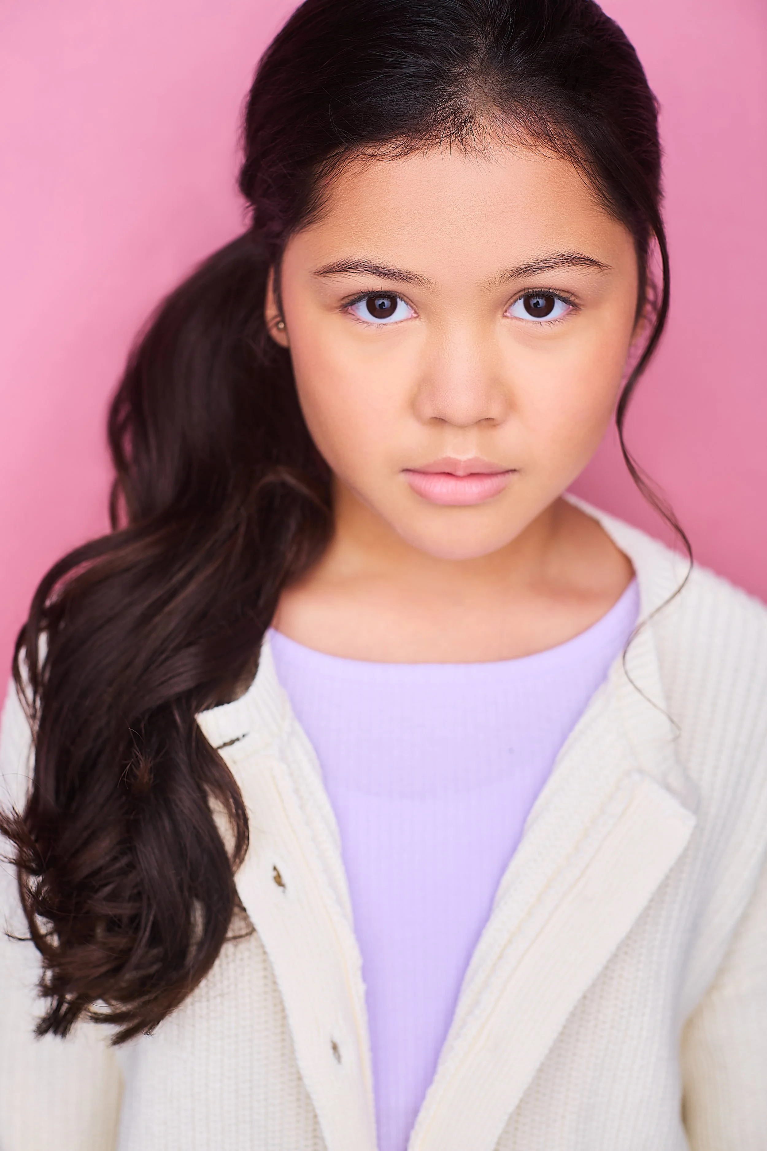 Close-up portrait of a young girl with long dark hair, wearing a lavender shirt and a cream cardigan, against a pink background.