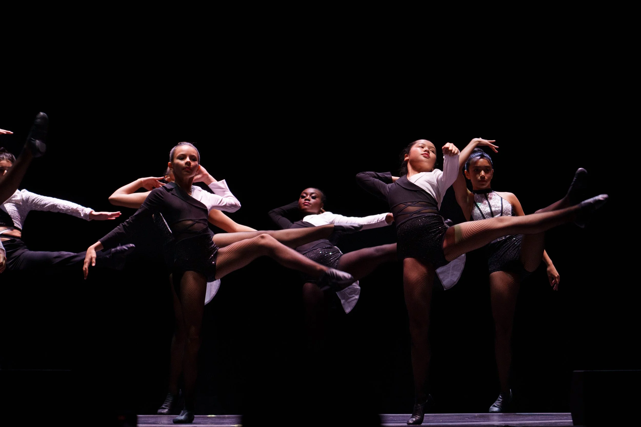 Group of female dancers performing a high kick on stage with black background.