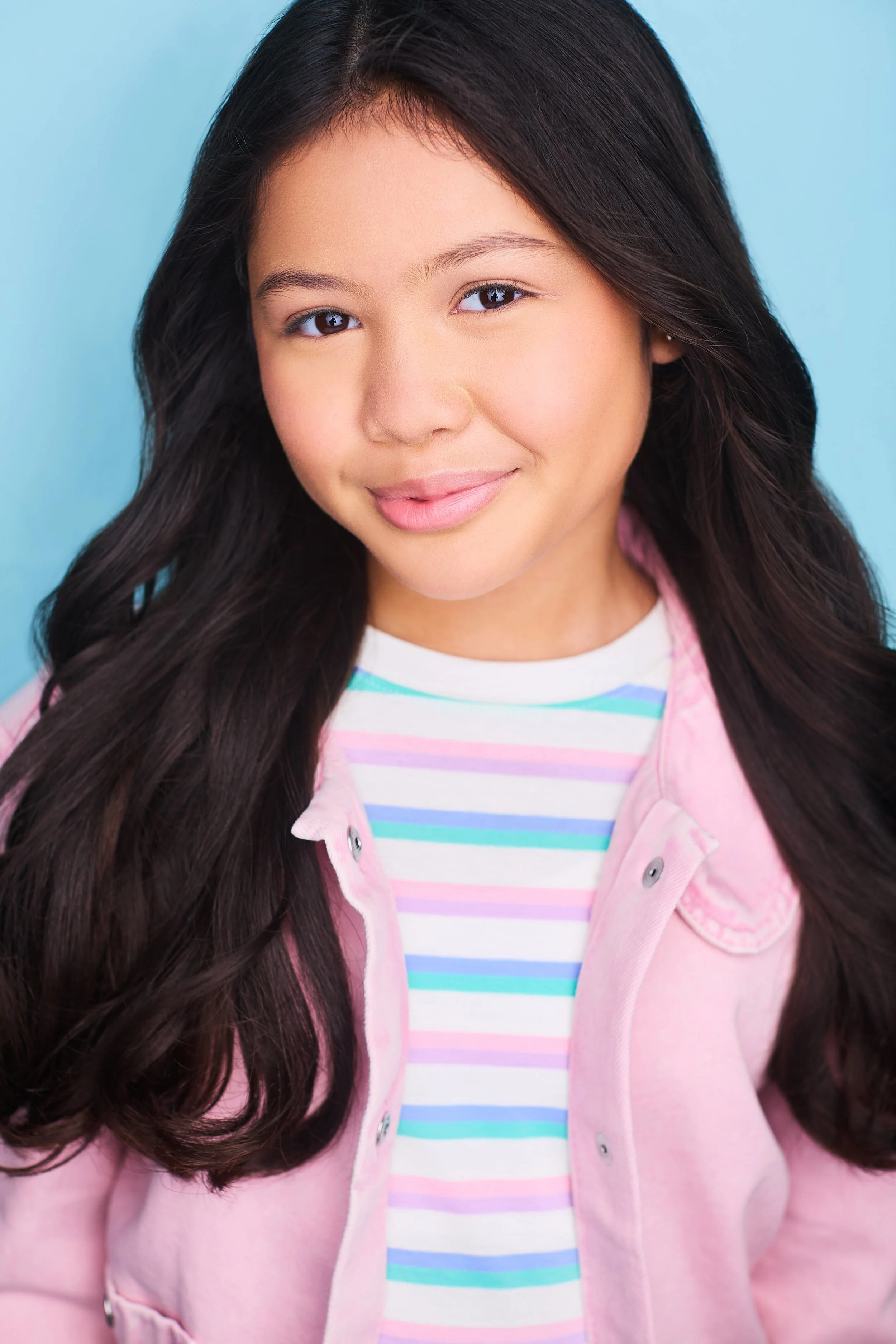 Young girl with long wavy dark hair, wearing a striped t-shirt and pink jacket, smiling against a blue background.
