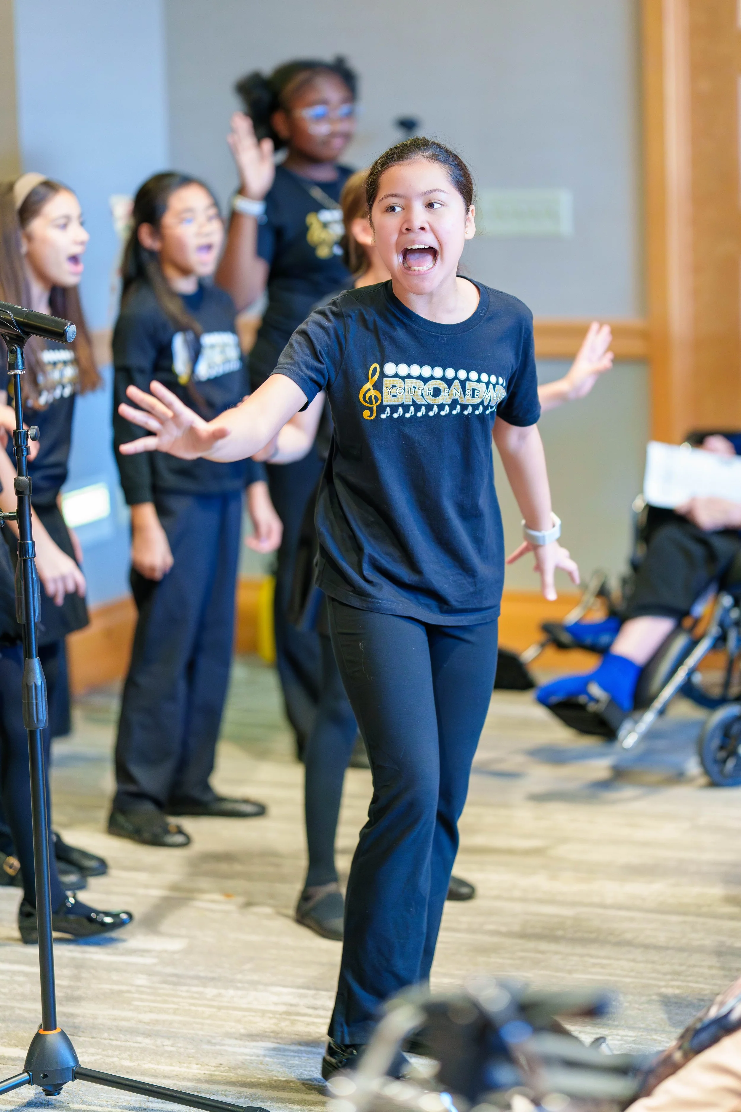 Young girl with dark hair singing passionately in front of a group of children during a choir practice or performance, wearing a navy T-shirt with music notes and the word 'Youth' on it.